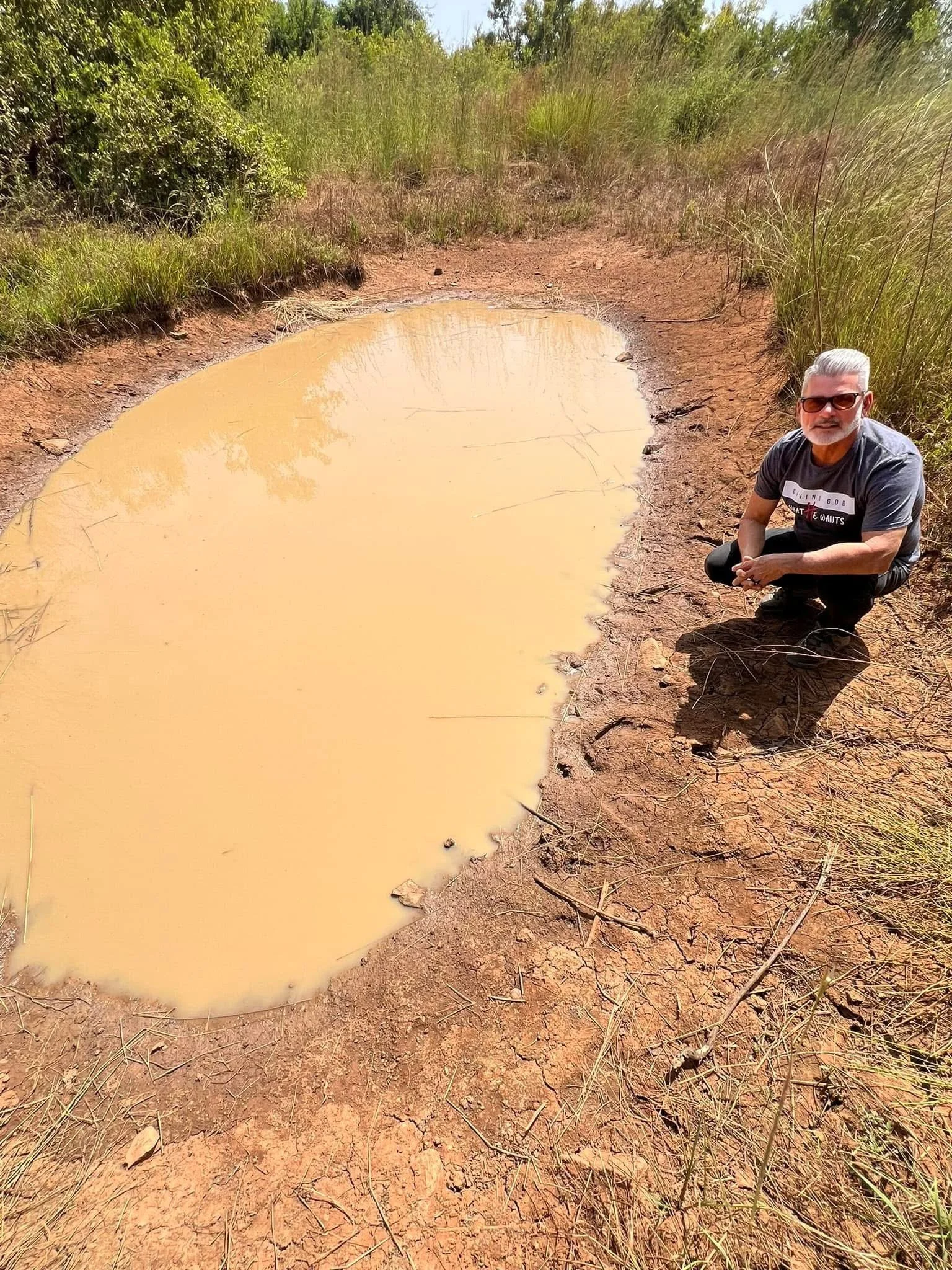A man wearing sunglasses and a gray T-shirt crouching beside a muddy waterhole in a dry, grassy area with green bushes in the background.