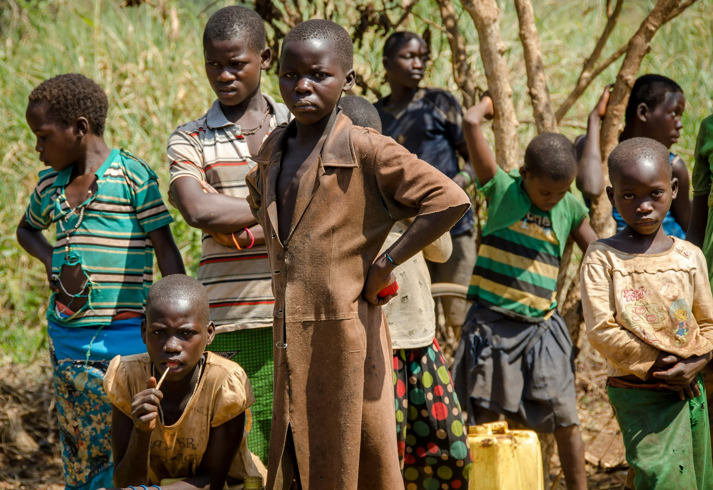 A group of children standing outdoors in a rural area, some with arms crossed, amidst trees and grass.