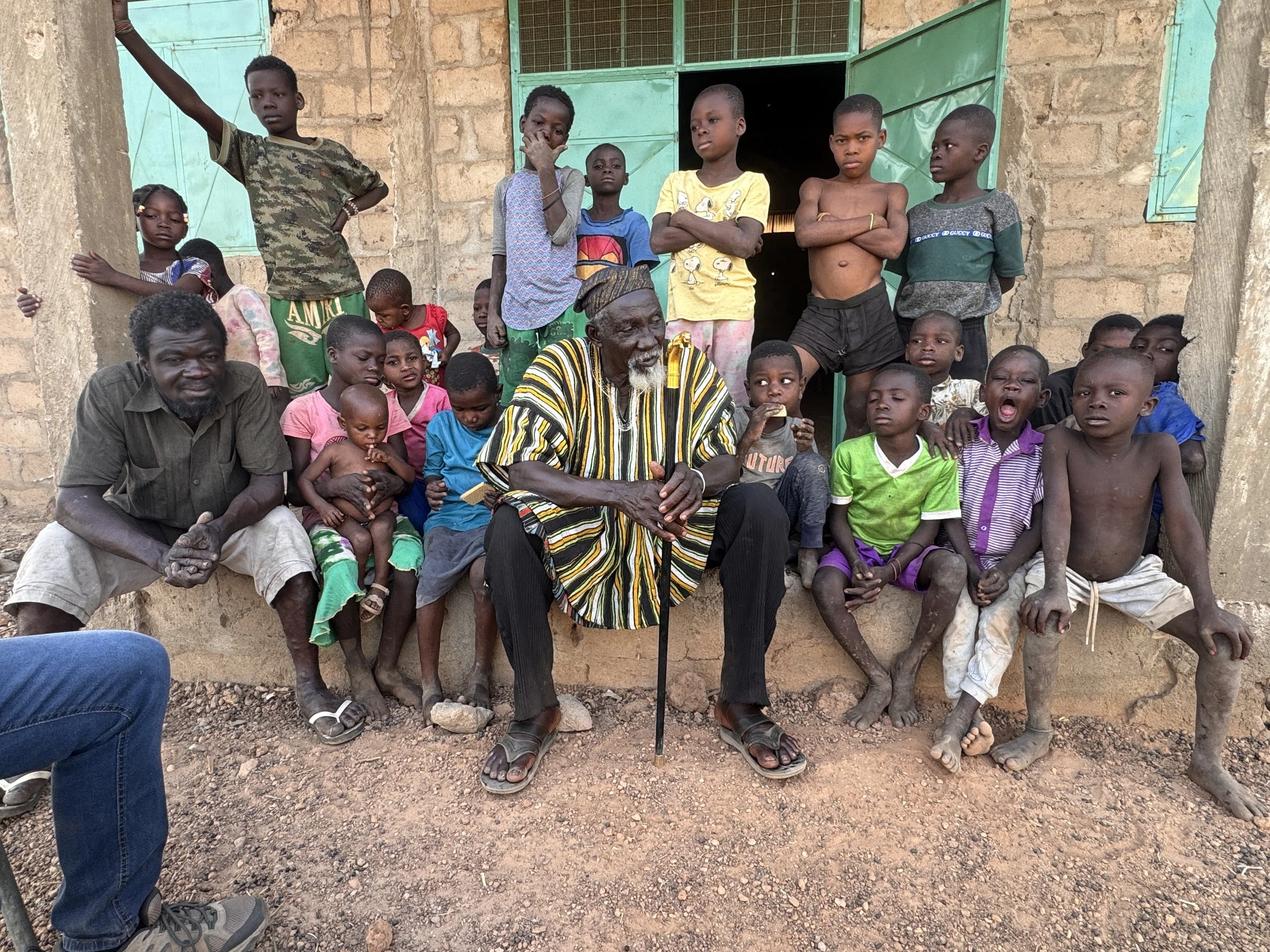A group of children and two adults sitting and standing outside a stone building with green door and window shutters. The children look serious or curious, some are barefoot or wearing sandals.