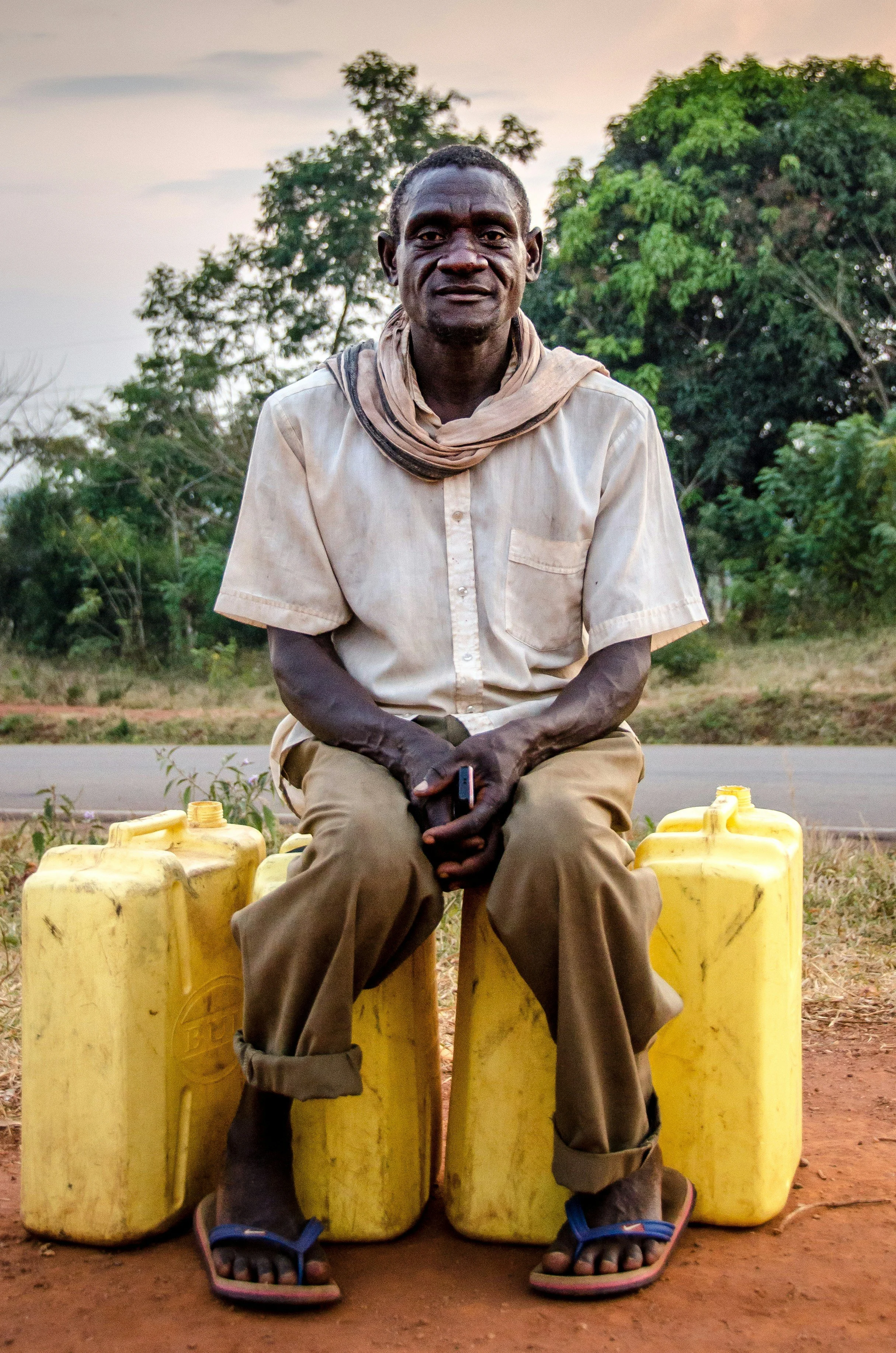 A man with dark skin, wearing a beige shirt with rolled-up sleeves, brown pants, and flip-flops, sitting outdoors on plastic containers with a backdrop of trees and a pastel-colored sky.