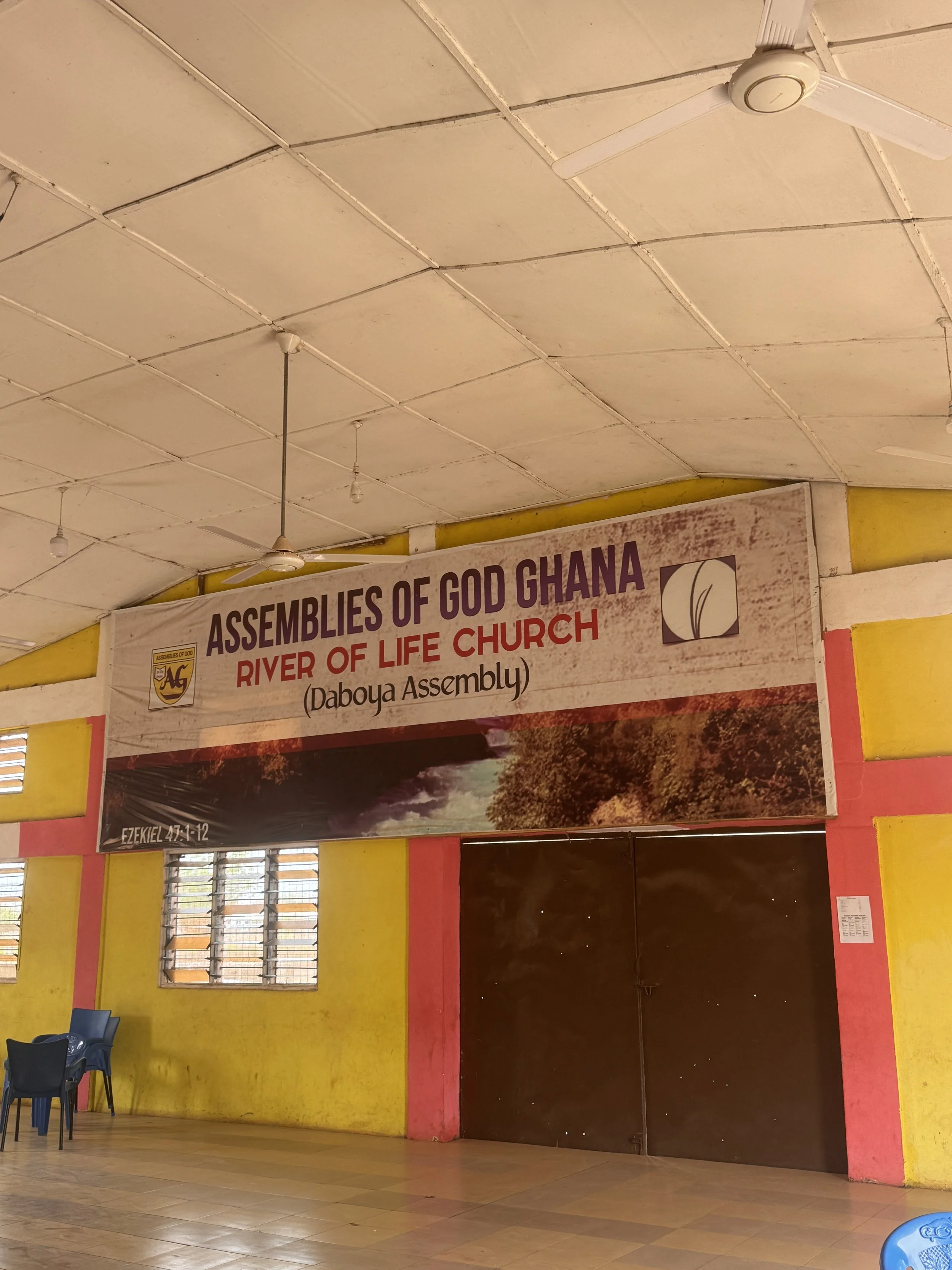 Interior of a church with a yellow and pink painted wall and a banner hanging that reads "Assemblies of God Ghana, River of Life Church, Daboya Assembly," with a view of windows, chairs, and a ceiling with ceiling fans and bulbs.