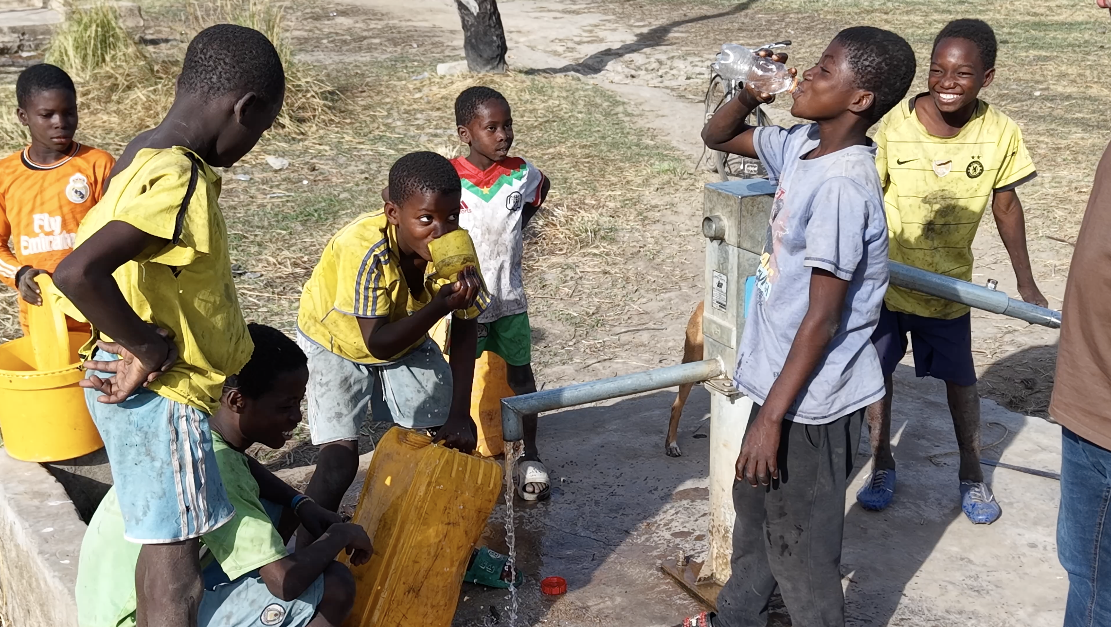 A group of children gather around a water pump, with some drinking water and others waiting. They are outdoors in a dry, dusty area.