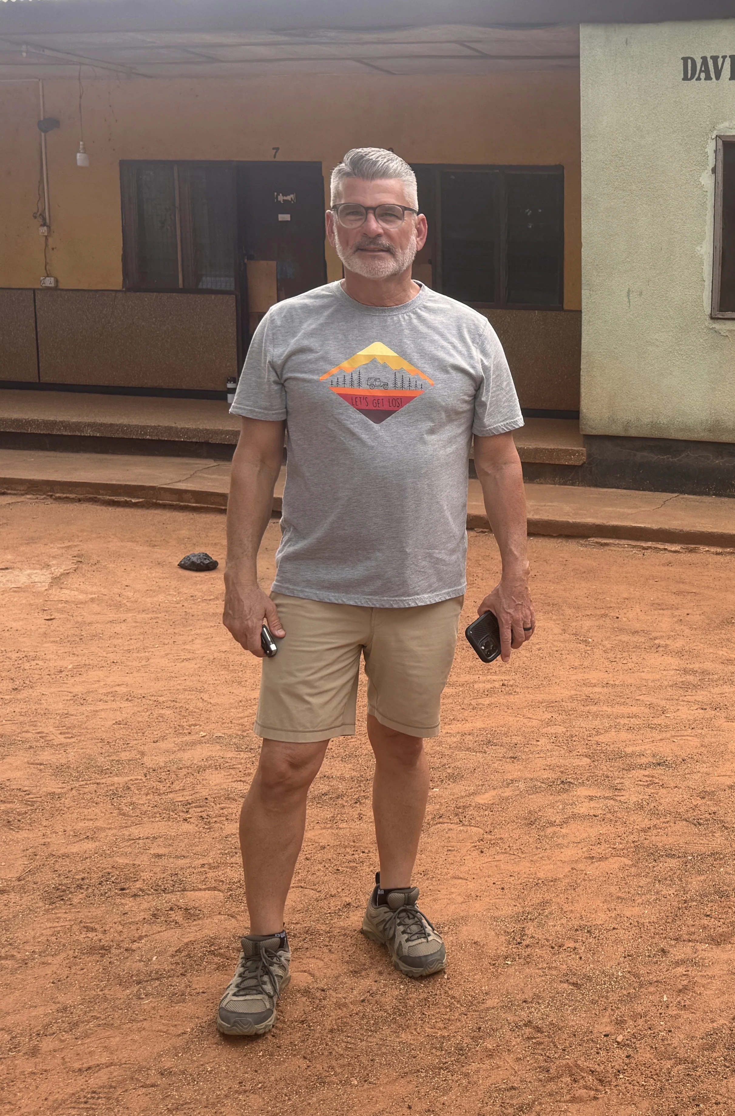 A middle-aged man with gray hair, glasses, and a beard standing on a dirt path outside a building, holding a phone in his right hand and wearing a gray T-shirt with a mountain graphic and the words "Let's Get Lost," khaki shorts, and hiking boots.