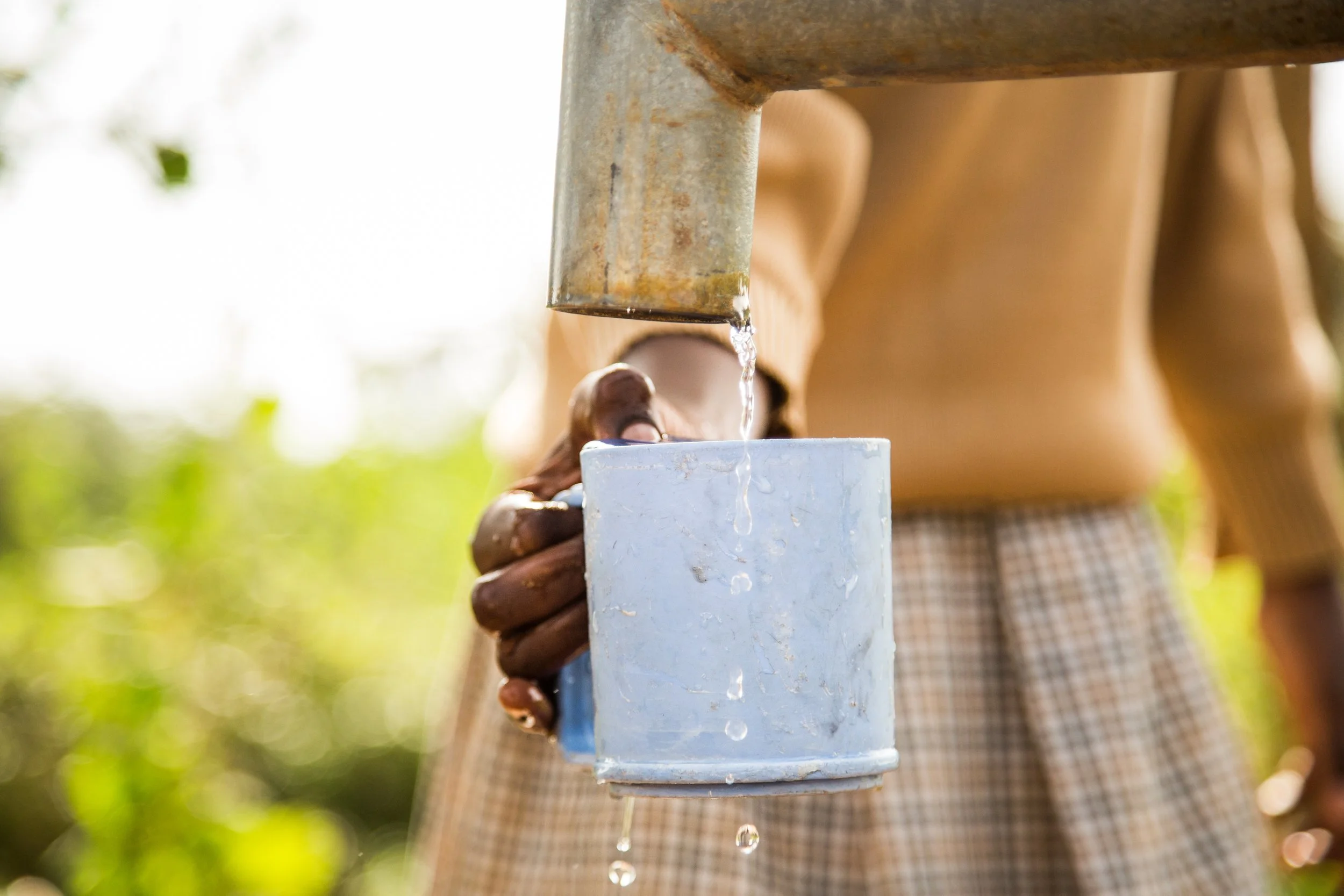 A person is pouring water from a metal spigot into a white container outdoors with greenery in the background.