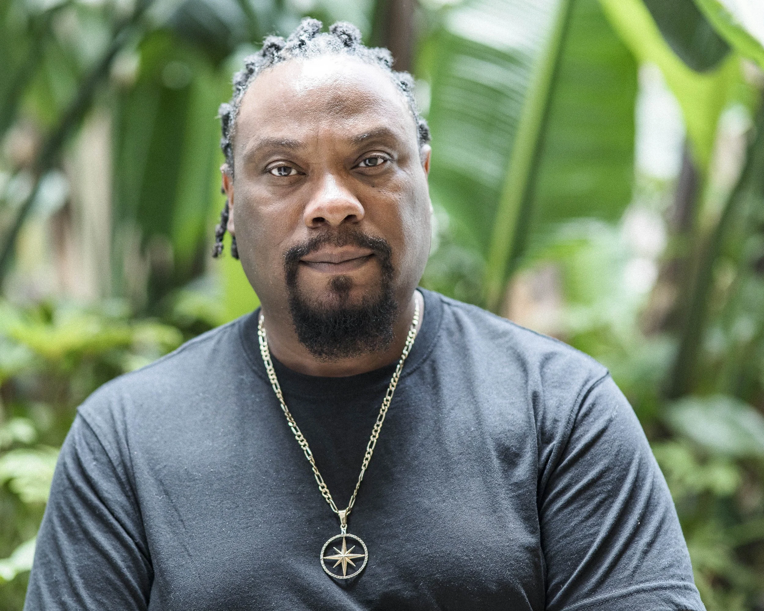 Close-up of Robin North with braided locs hair, a mustache, and a goatee, wearing a black t-shirt and a gold necklace with a pendant of Polaris or North Star, a custom-made necklace in honor of his grandmother, Susie North.