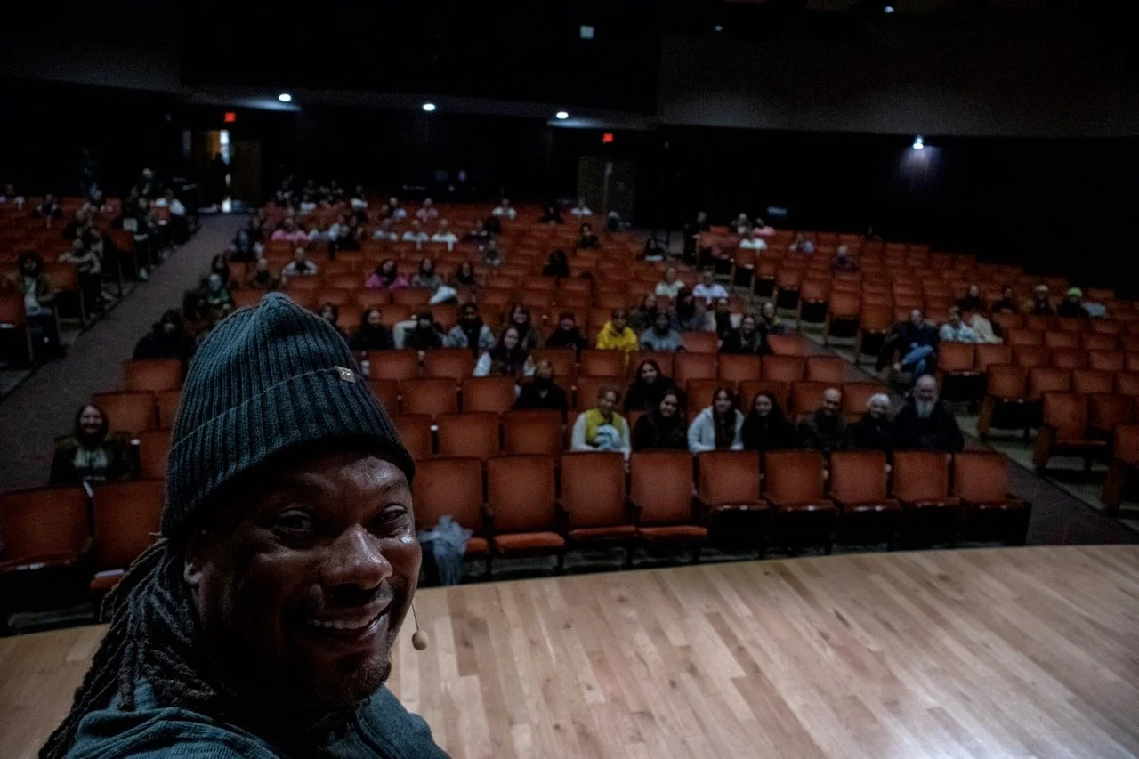 Robin North is composing an image with the attendees of his Artist Talk at NW Missouri State University with a microphone, overlooking an audience seated in an auditorium.