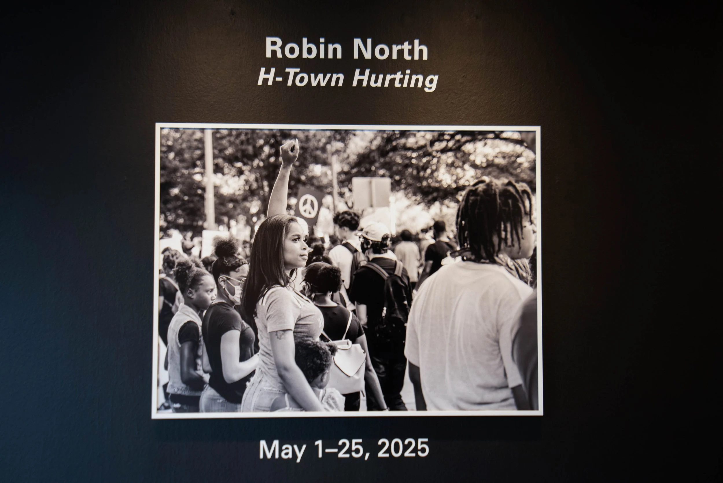 The image shows a  photograph of a group of people participating in a protest. It features a woman with long hair and a face mask, and a person with dreadlocks. They are among other participants holding signs and banners. The text on the display indicates the event is titled "Robin North" with a subtitle "H-Town Hurting," scheduled for May 1-25, 2025.