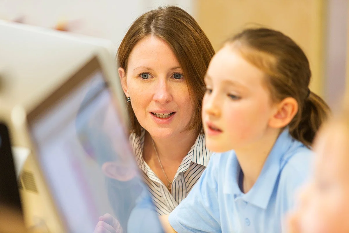 A female teacher sits beside a primary school age student as they work together on a computer.