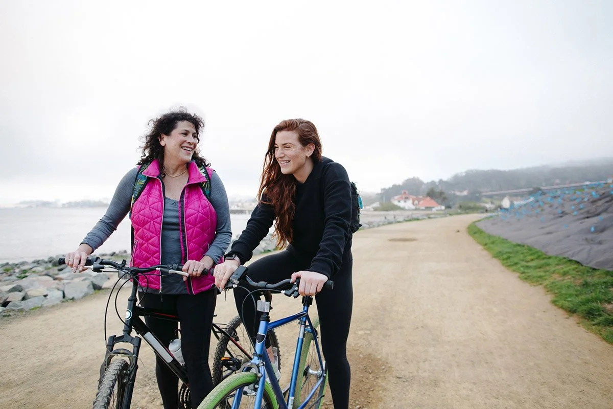 A middle age woman and young woman in early 20s resting with their bicycles near a rocky coastline.