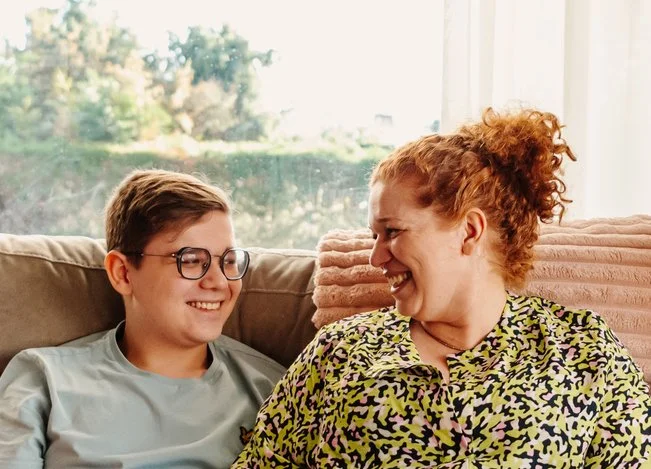 A red haired woman sits next to a smiling teenage male indoors, with a view to a garden out the window