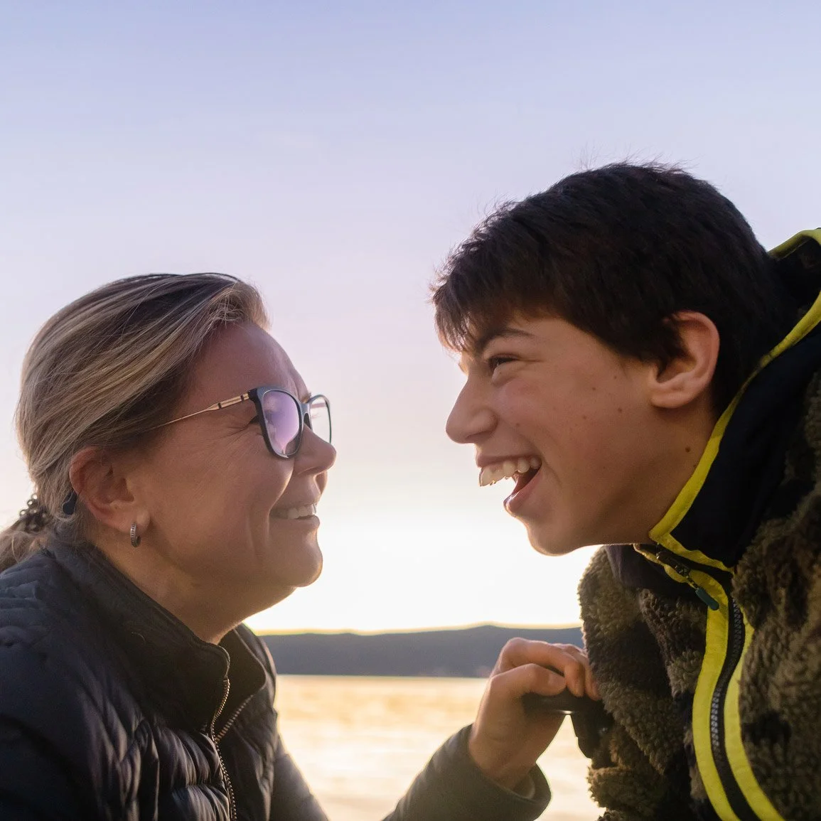 A mother and teenage son facing eachother, smiling and laughing, against a sunset sky.
