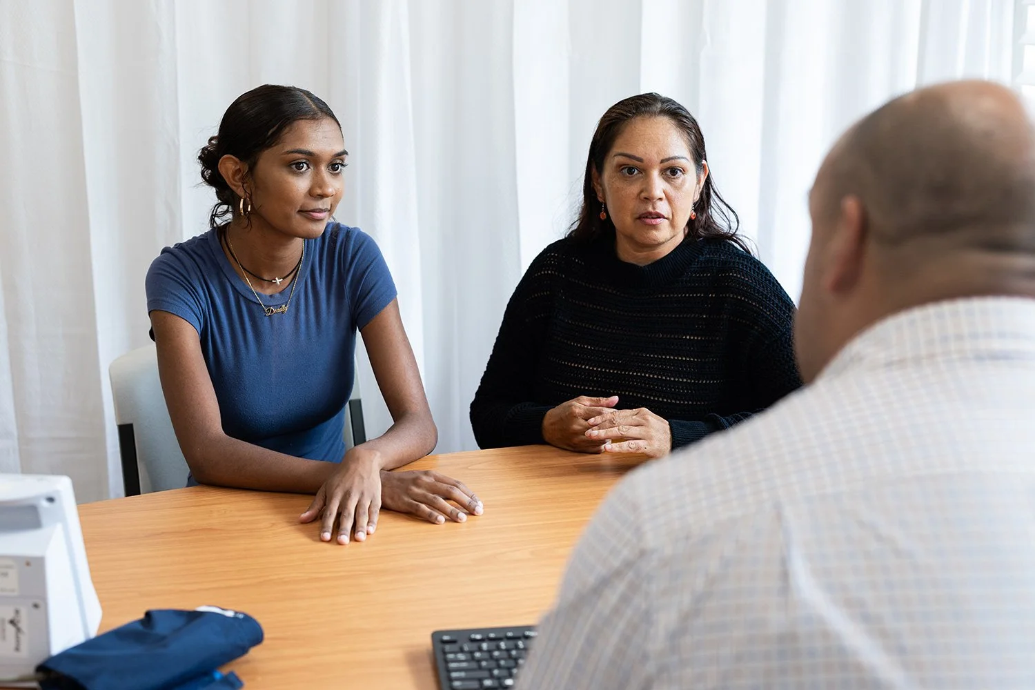 Two women sitting across from a man at a wooden table in a professional setting, engaged in conversation. The women are looking attentively at the man, who is partially visible and facing away from the camera.