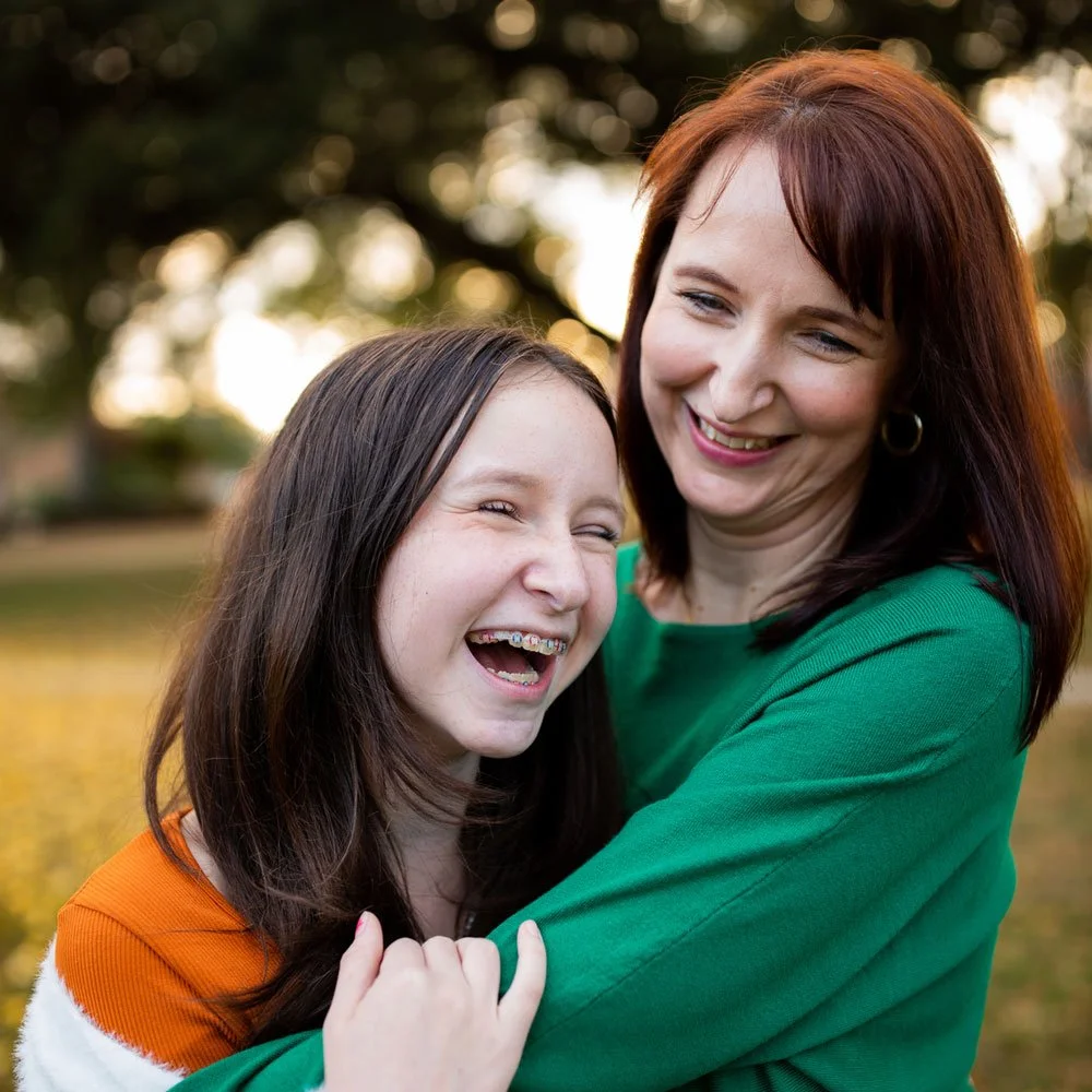 A smiling mother and daughter with long dark hair embrace in an outdoor setting