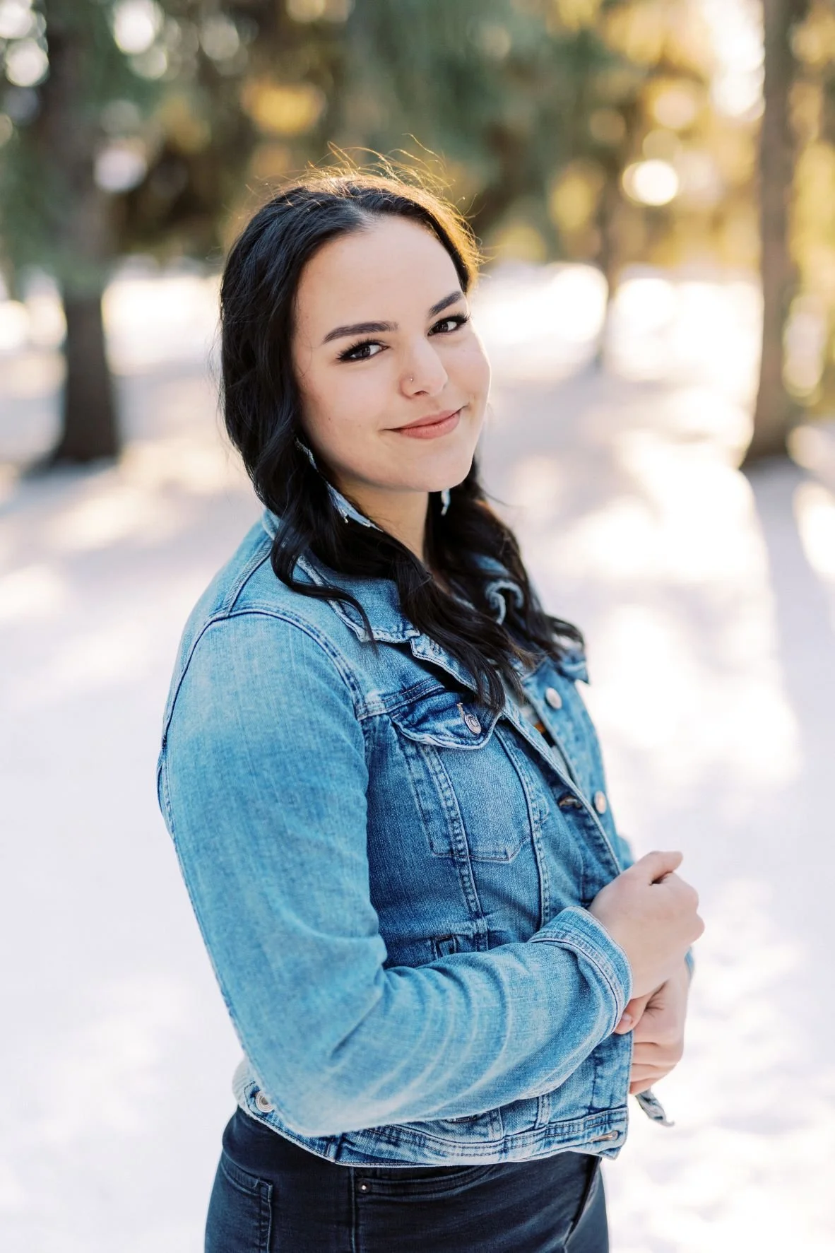 A young woman with dark hair and a nose piercing, wearing a blue denim jacket, stands outdoors in a snowy environment with trees in the background, during sunset or golden hour.