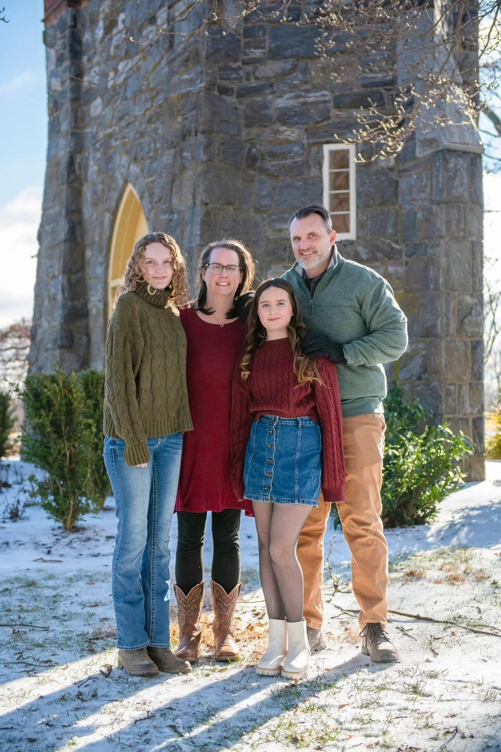 A family of four standing outside on a snowy day in front of a stone building with a small window and trees. The family members are dressed in warm clothing and smiling at the camera.