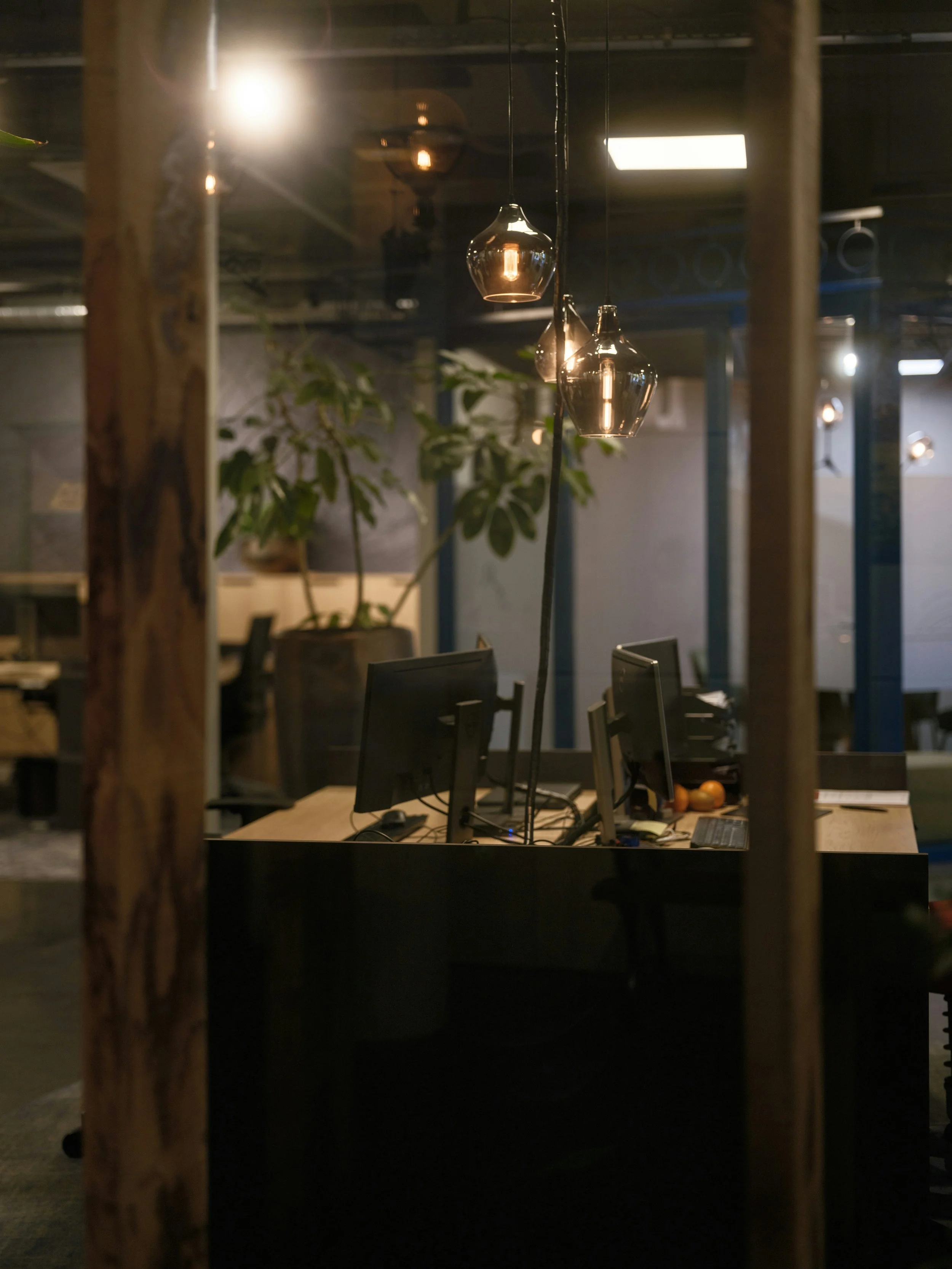 Dimly lit office space with multiple computer monitors, a wooden desk, and hanging pendant lights, viewed through a glass partition.