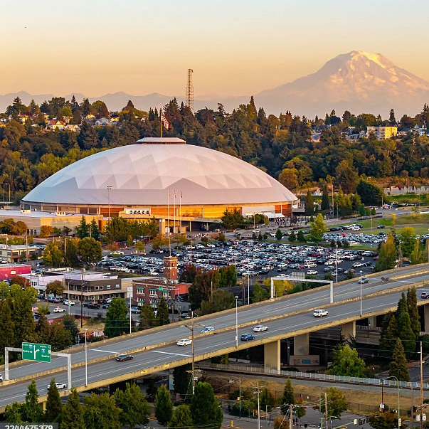 A large white dome building with parking lot, surrounding trees, and a highway in front. Mountain in the background at sunset.