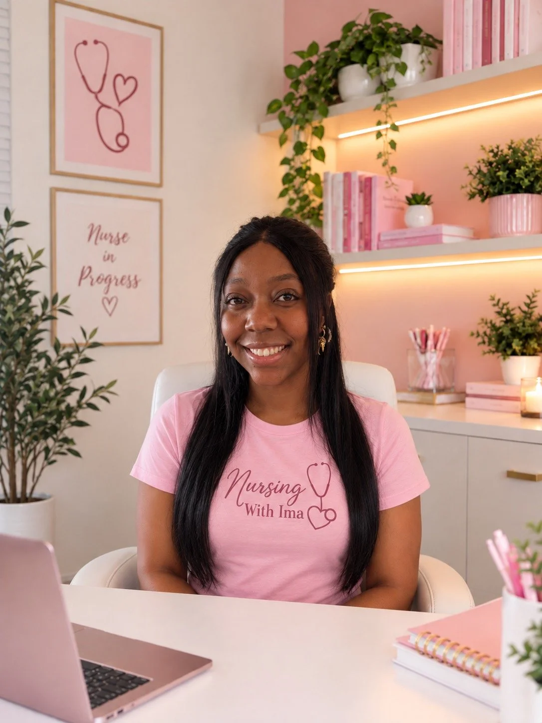 A woman smiling and sitting at a desk with a pink shirt that says 'Nursing With Ima' along with a stethoscope graphic. The background is decorated with pink and white elements, including framed art that reads 'Nurse in Progress', shelves with books, potted plants, and pink accents.