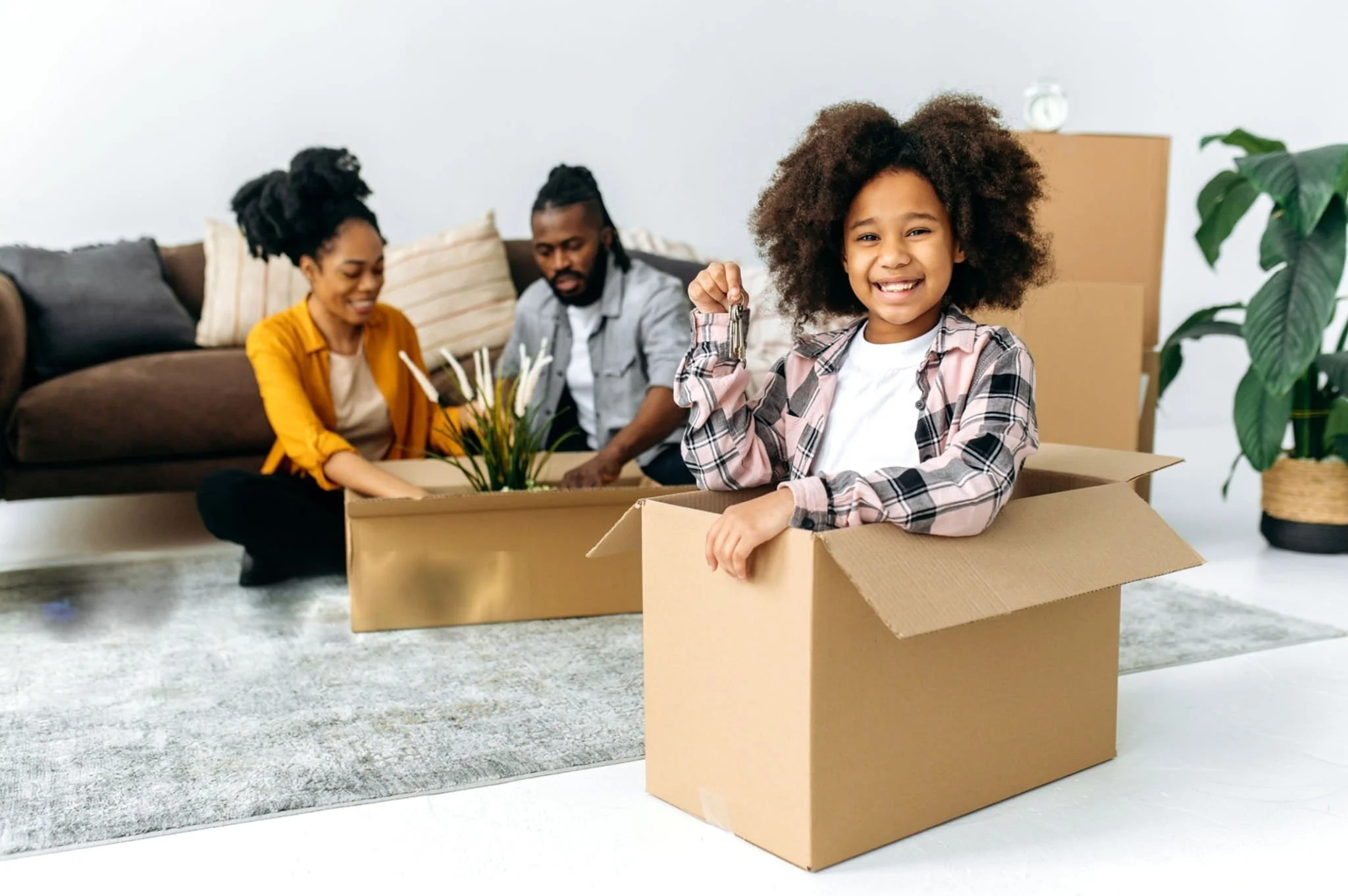 Une famille de trois personnes, un père, une mère et une fille, s'apprête à déménager dans leur nouveau logement. La fille, qui a de gros cheveux afro, tient une clé dans sa main et sourit, assise dans une boîte en carton. La mère et le père sont assis derrière elle sur un canapé, discutant ou organisant des affaires, avec des cartons de déménagement autour d'eux.