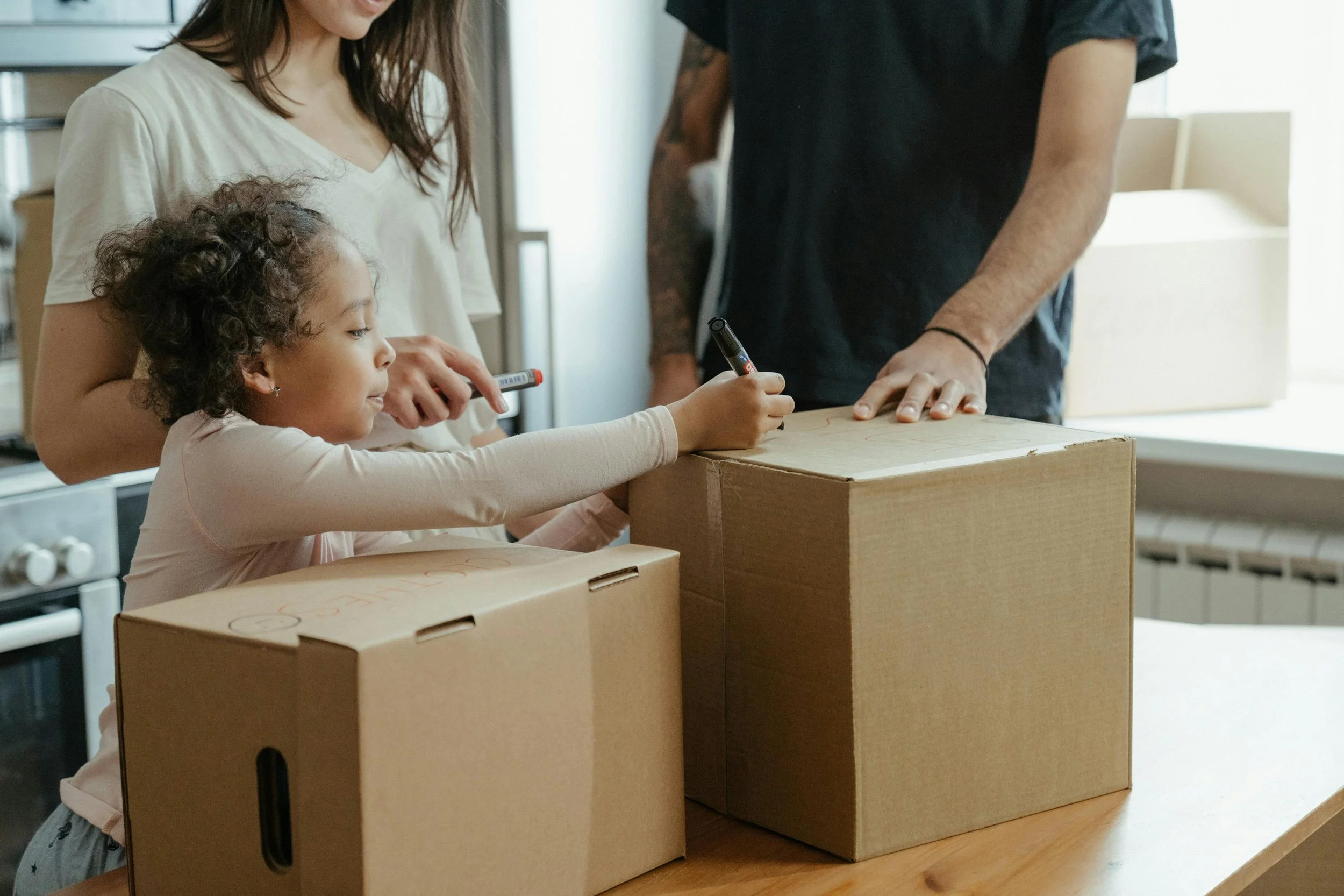Une jeune fille signe un contrat avec un adulte dans un environnement intérieur, entourée de trois cartons en carton kraft sur une surface en bois.