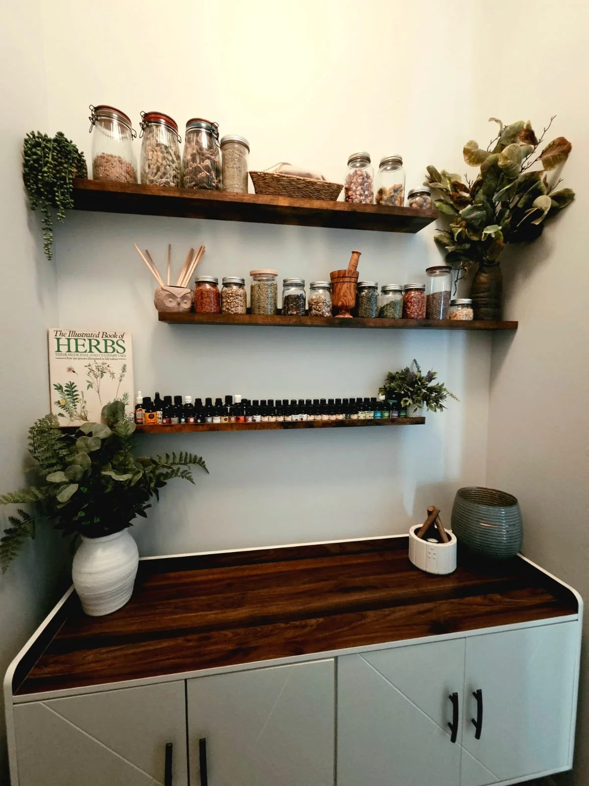 Three rustic wooden floating shelves displaying glass jars of dried herbs, botanical ingredients, amber essential oil bottles, and a book on traditional herbs above a dark wood counter.