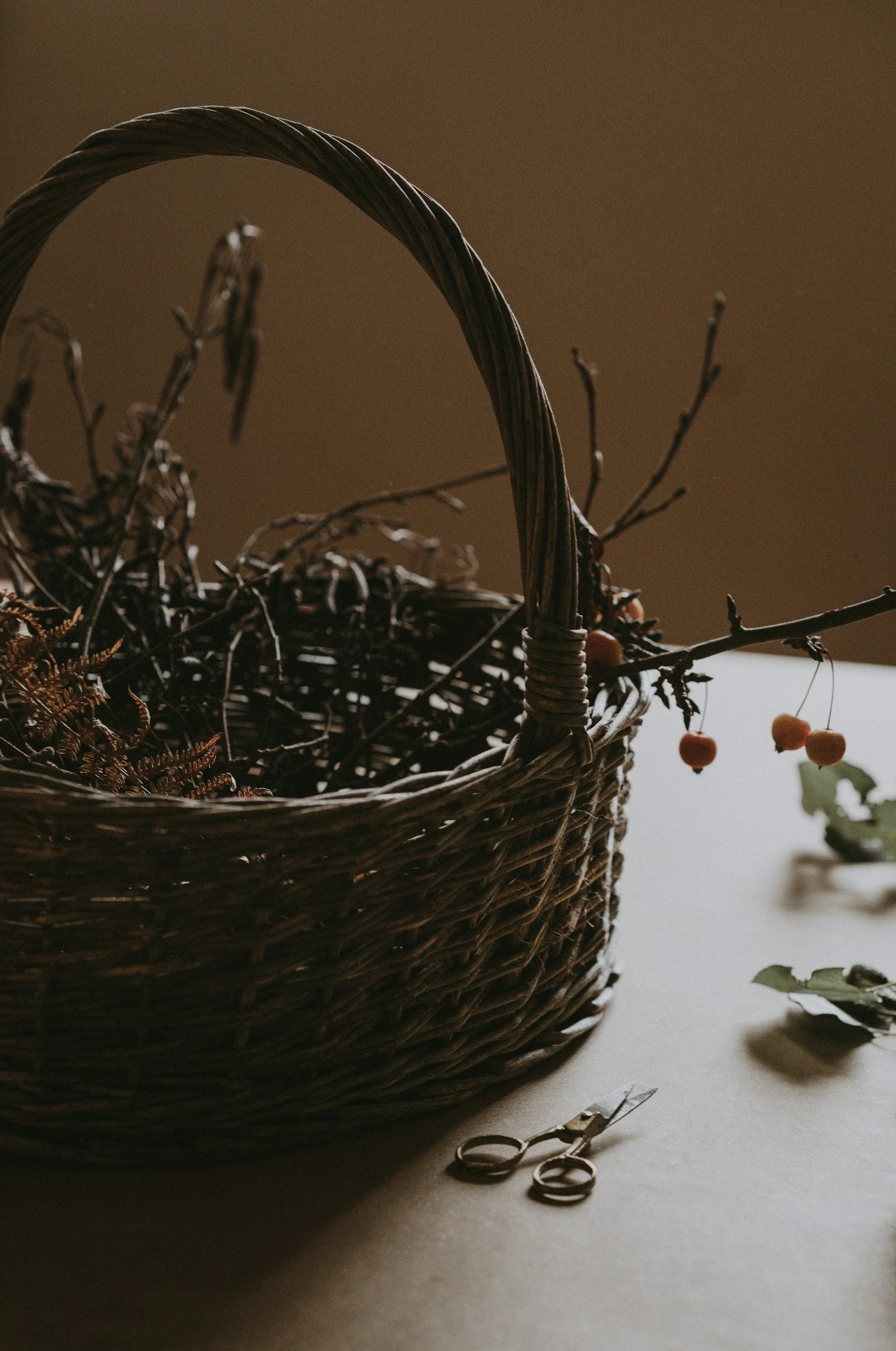 A woven wicker basket with dried plants or flowers inside, on a table with a pair of scissors nearby, and a small potted plant with red berries in the background.