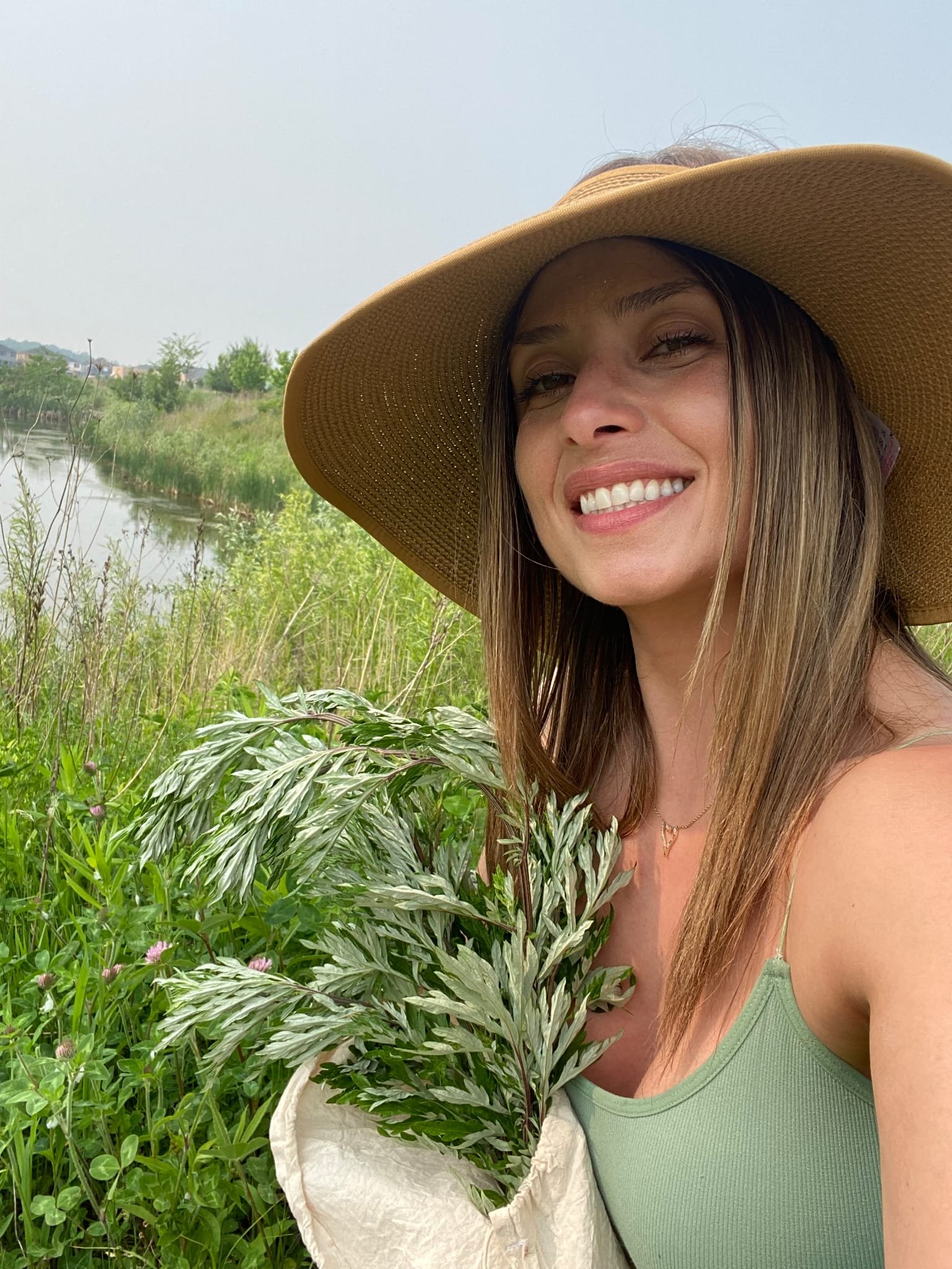 A woman outdoors near a body of water, wearing a wide-brimmed hat and a green tank top, smiling and holding lush green and variegated plants.
