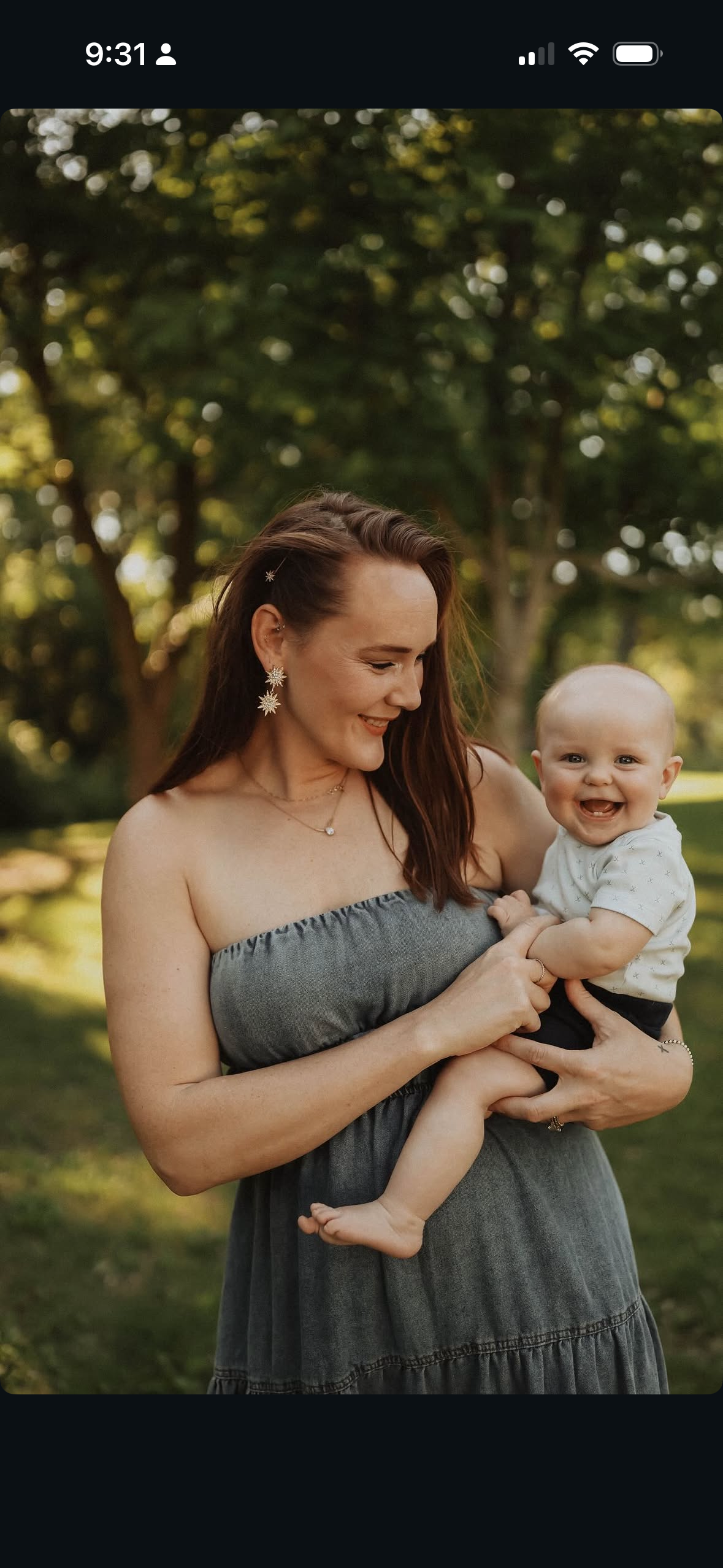 A smiling woman in a strapless gray dress holding a happy baby boy outdoors with trees in the background.