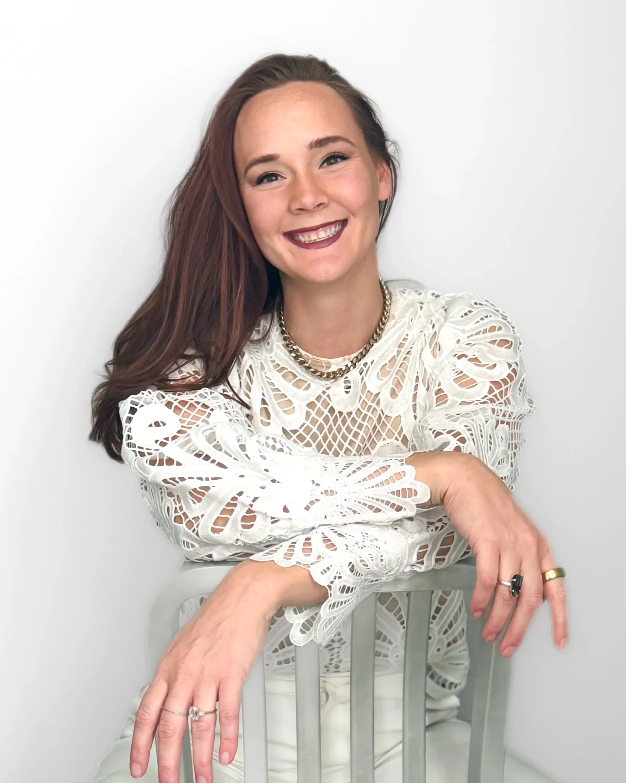 A young woman with auburn hair smiling, sitting on a modern silver chair against a plain white background, wearing a white lace top, gold necklaces, and rings.