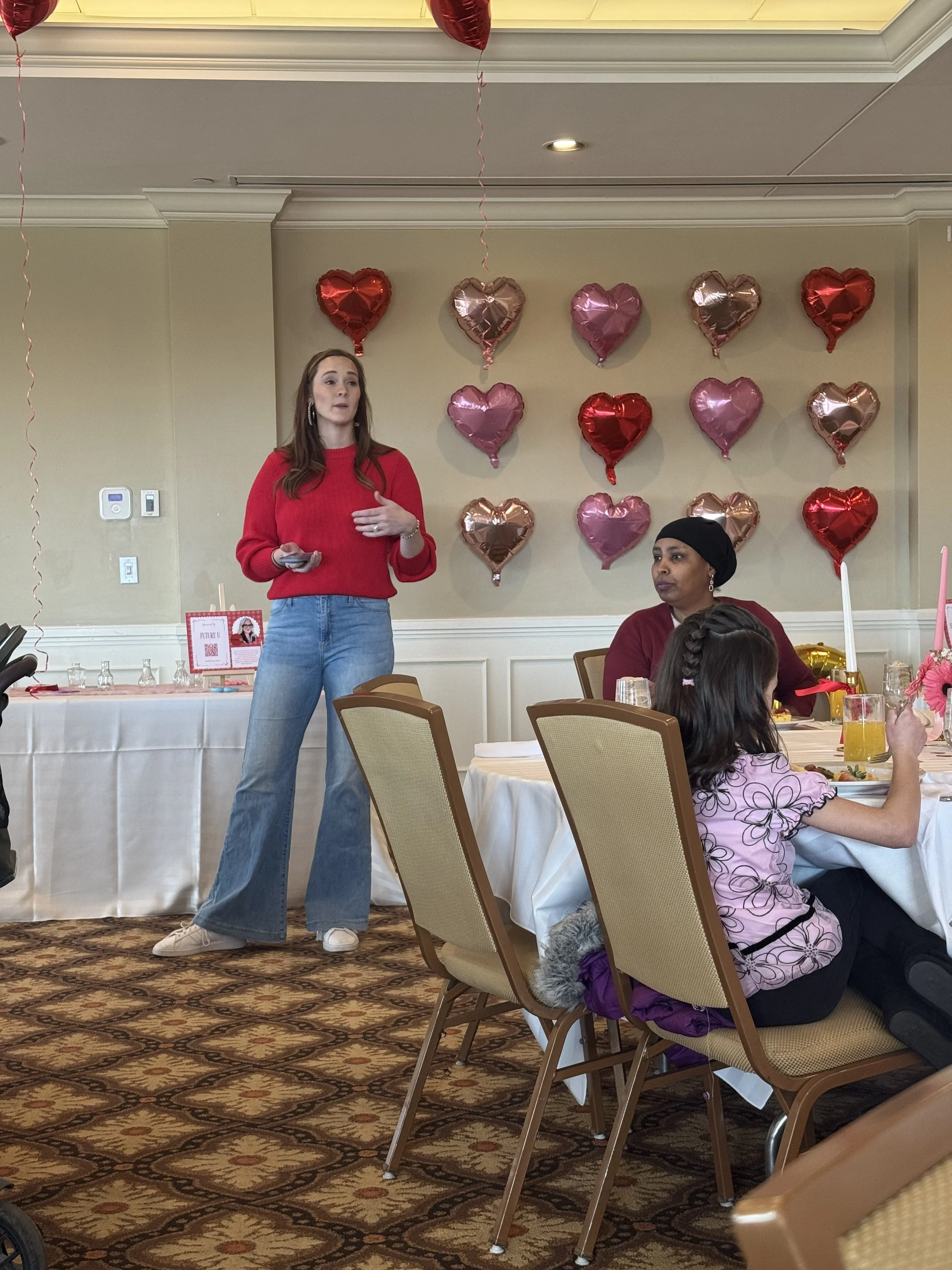 A woman in a red sweater and blue jeans speaking at a celebration event decorated with heart-shaped balloons in red and pink, with a woman sitting at a table with children in the foreground.