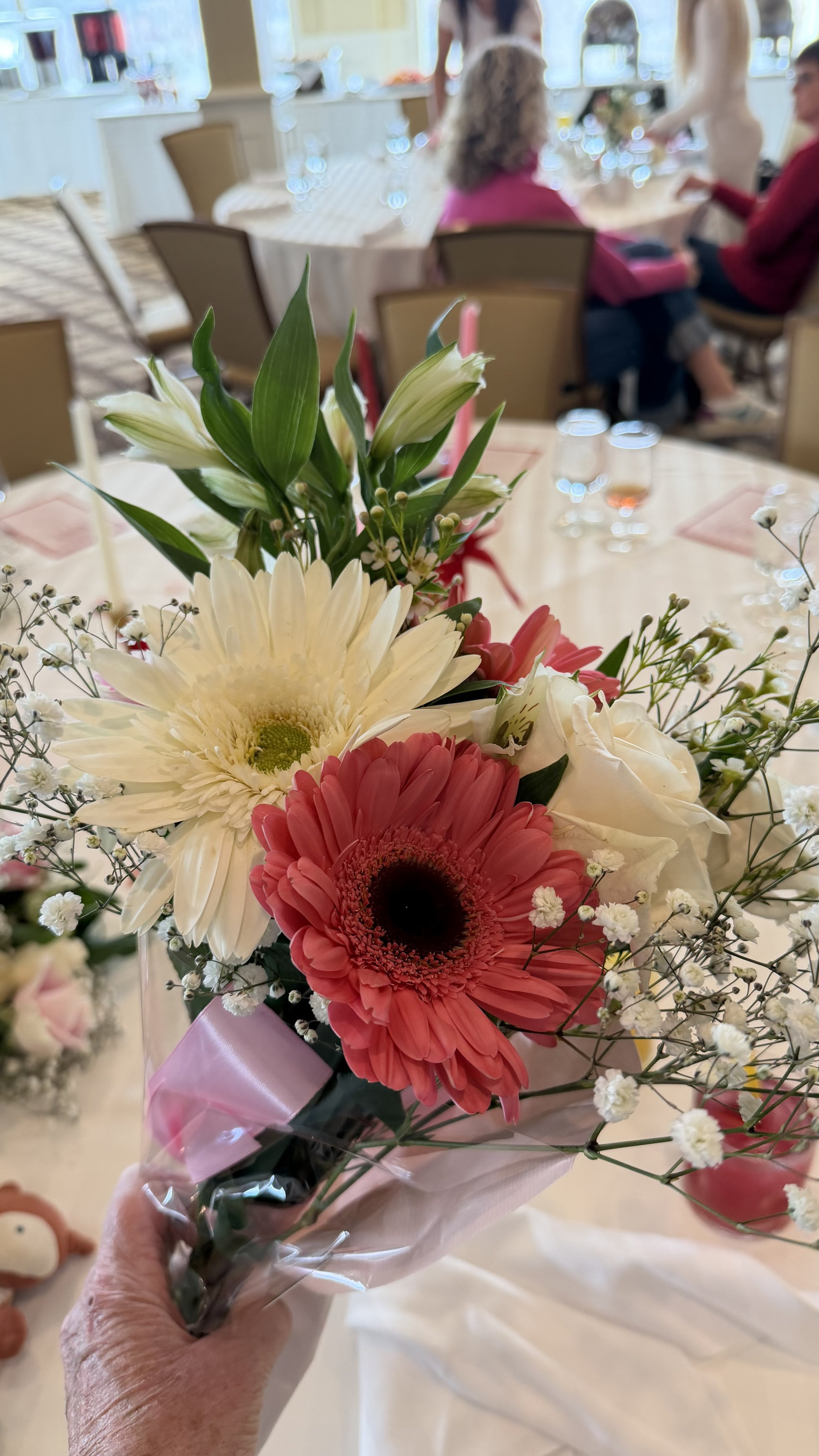 A bouquet of white, pink, and red flowers with green leaves, wrapped in pink and clear paper, held by a person's hand in a decorated dining room with people sitting at tables in the background.