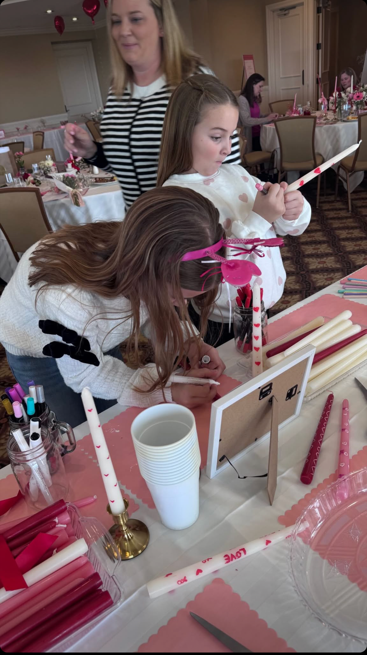 A young girl is writing on a paper at a decorated party table with pink and red items, including candles, cups, and paper straws with hearts. A woman stands nearby, and other girls are sitting at the background tables, all in a festive setting.