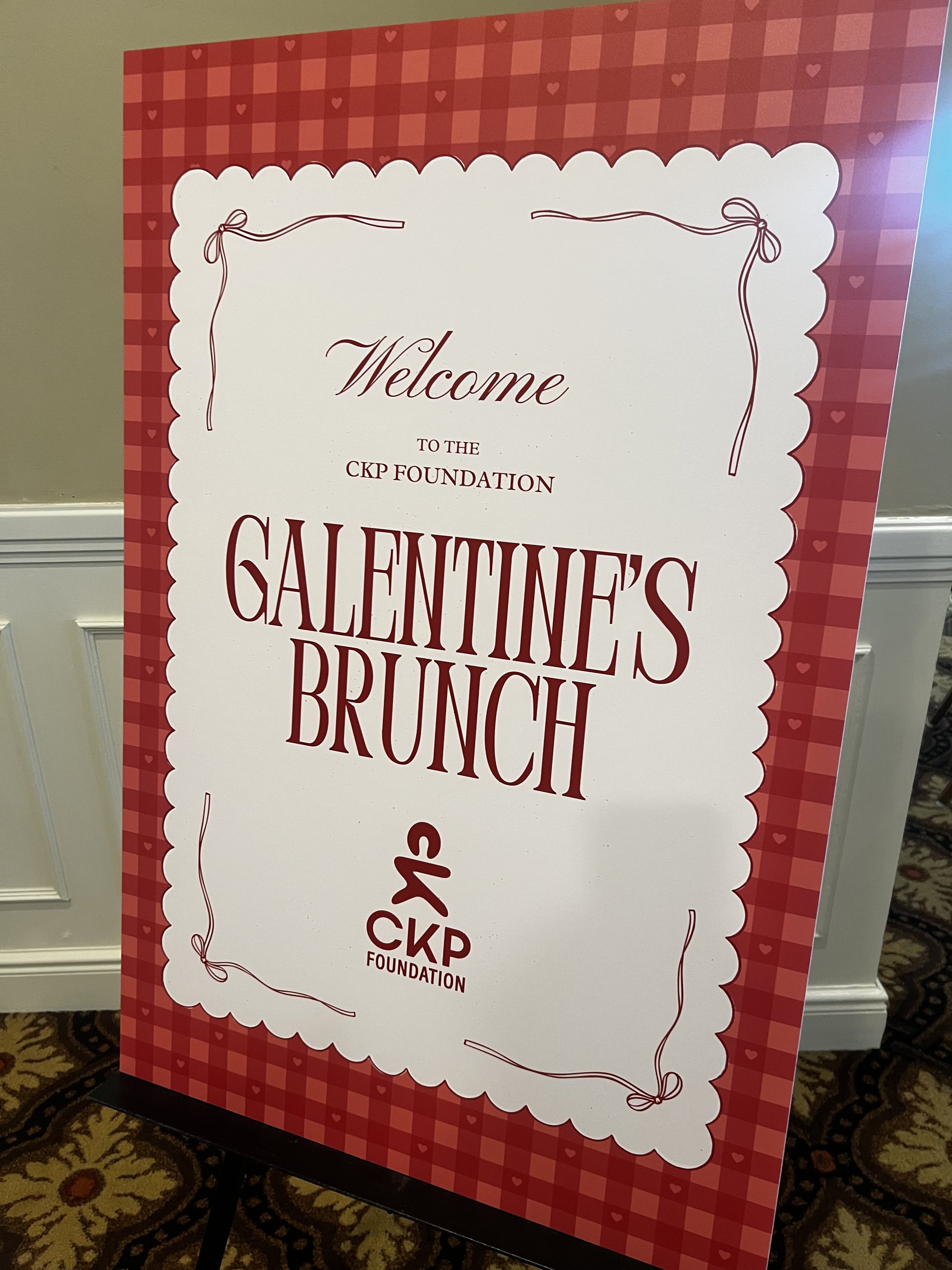 A decorative sign with a red buffalo plaid border, featuring the text 'Welcome to the CKP Foundation Valentine's Brunch' in elegant red and brown fonts. The sign has a scalloped edge and is decorated with small hearts and bow illustrations.