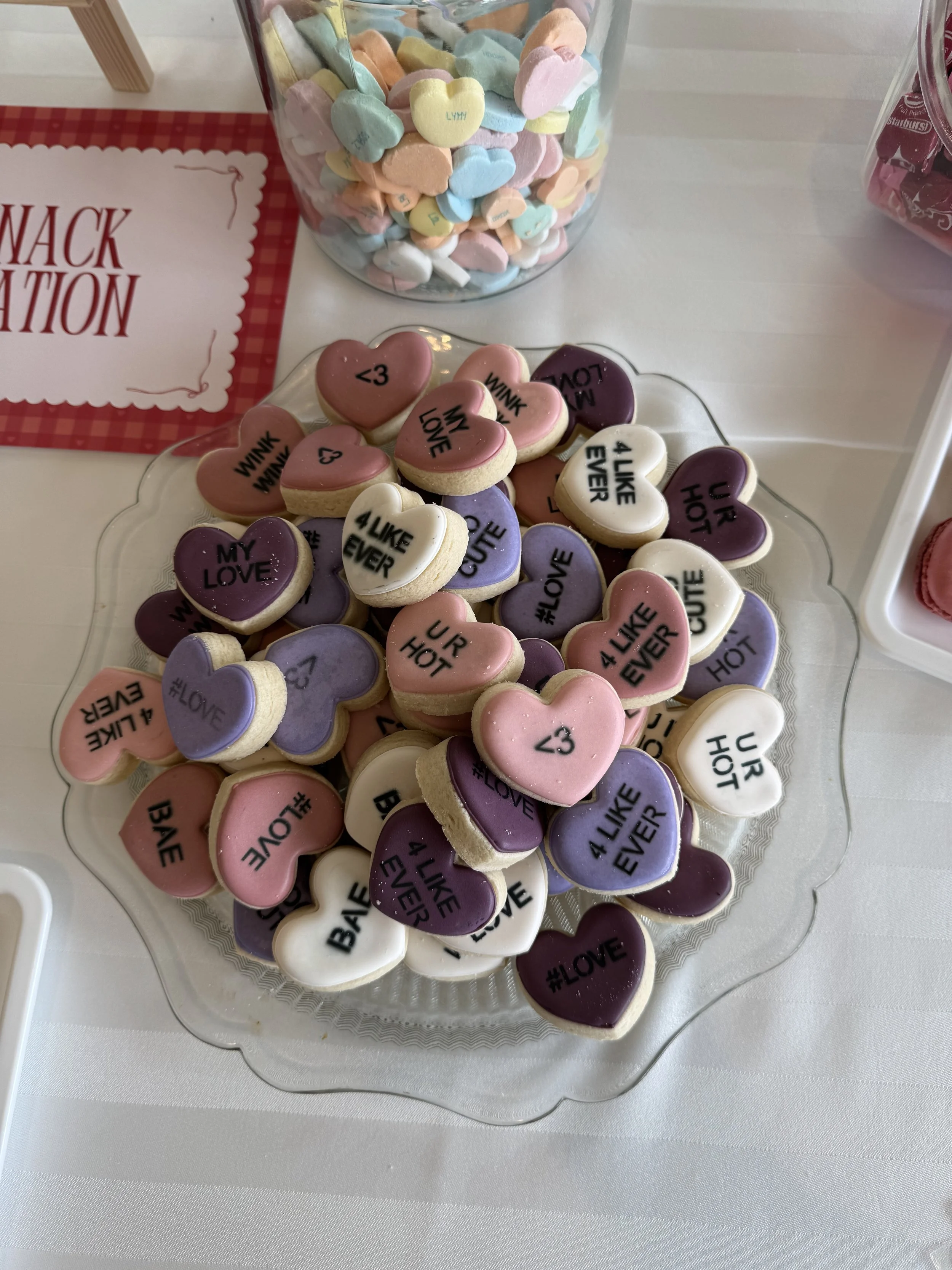 Heart-shaped cookies with pastel-colored icing and black messages such as 'LOVE,' '<3,' 'WINK WINK,' '#LOVE,' '4 U LIKE EVER,' and 'UR HOT,' arranged on a glass platter on a white table.