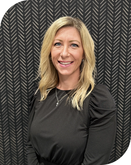 A woman with blonde hair smiling in front of a dark chevron-patterned background, wearing a black top and a cross necklace.