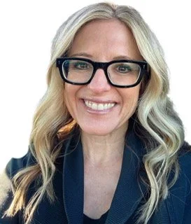 A woman with long blonde hair wearing black glasses and a dark blue blazer, smiling.