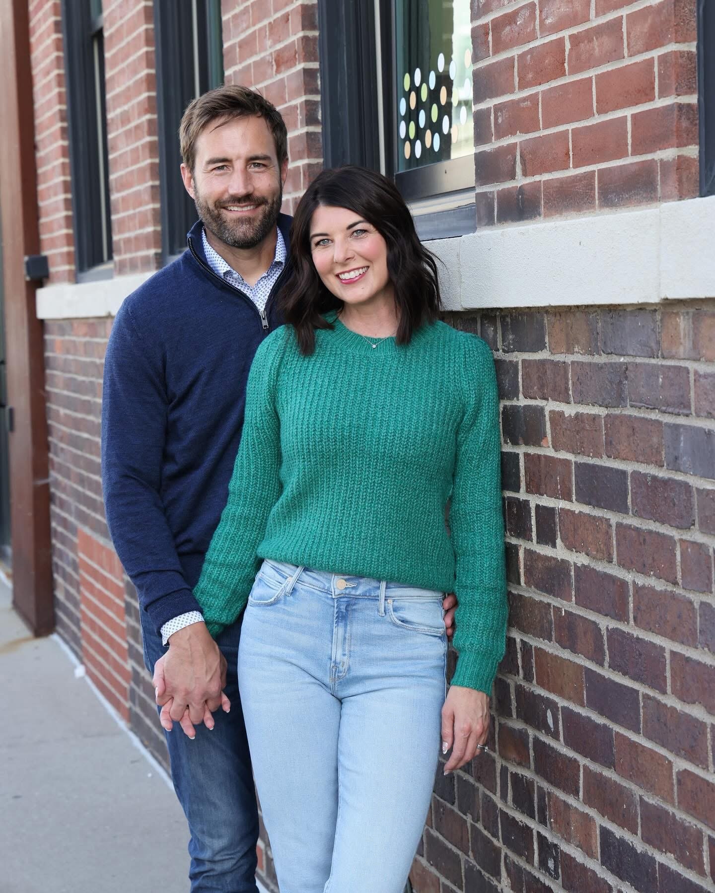 A smiling man and woman holding hands, standing against a brick wall on a sidewalk.