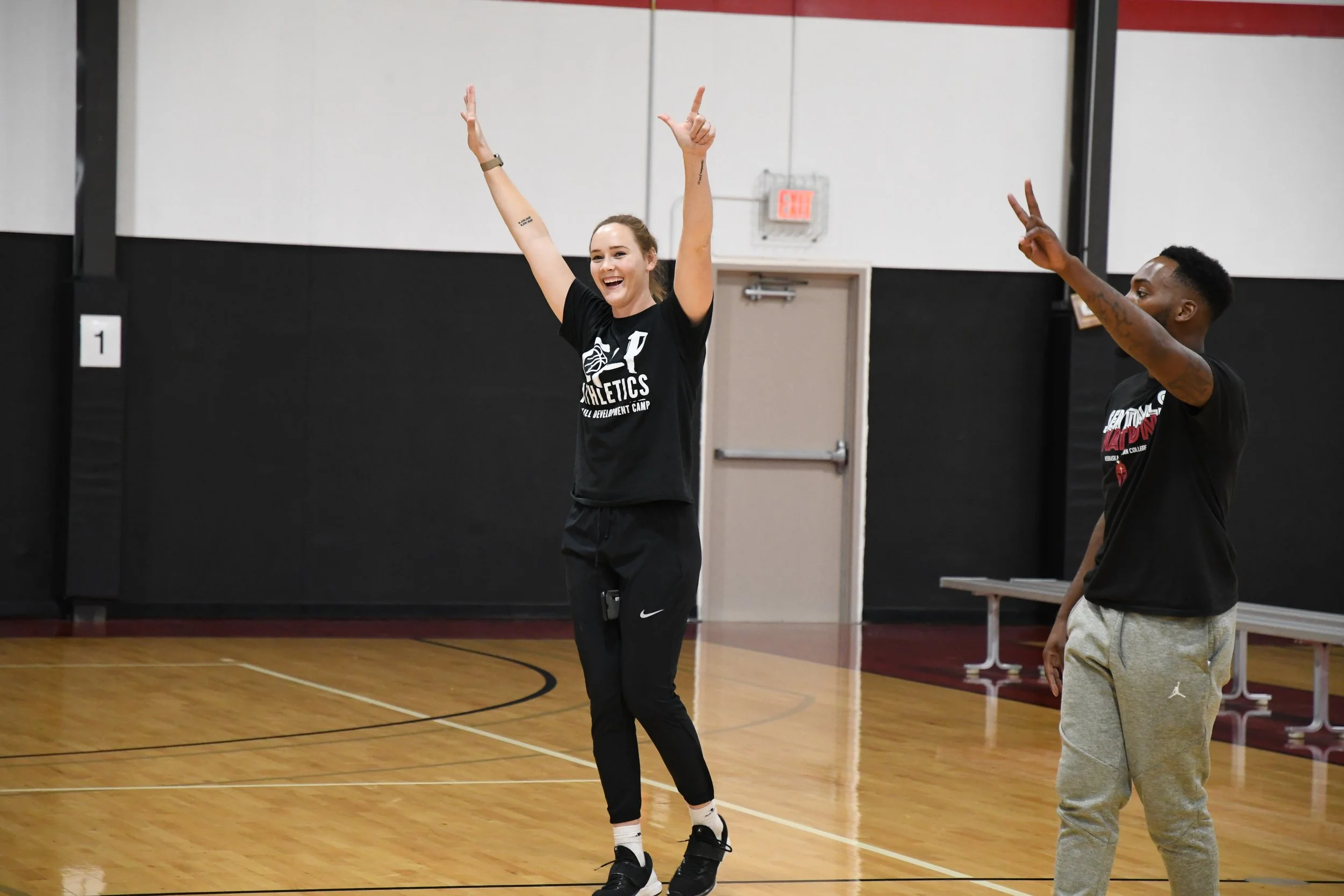 Two people on a basketball court, one woman with her arms raised and smiling, the other man making a peace sign, both wearing black T-shirts.