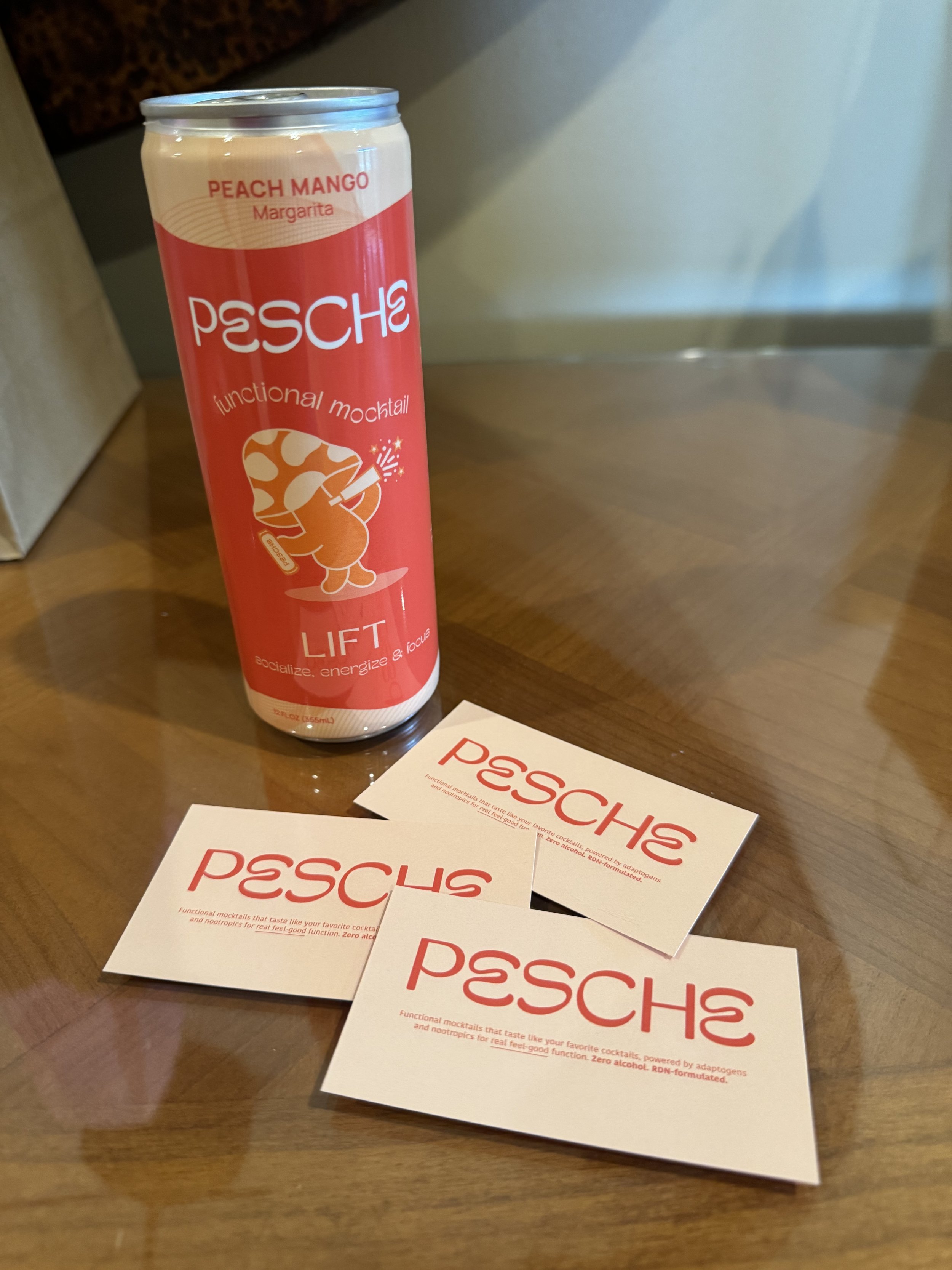 A can of Pesche Peach Mango Margarita standing on a wooden table with several business cards spreading out in front of it. The can is red with white text and a graphic of a mushroom wearing sunglasses, with the words 'functional mocktail' written around it.