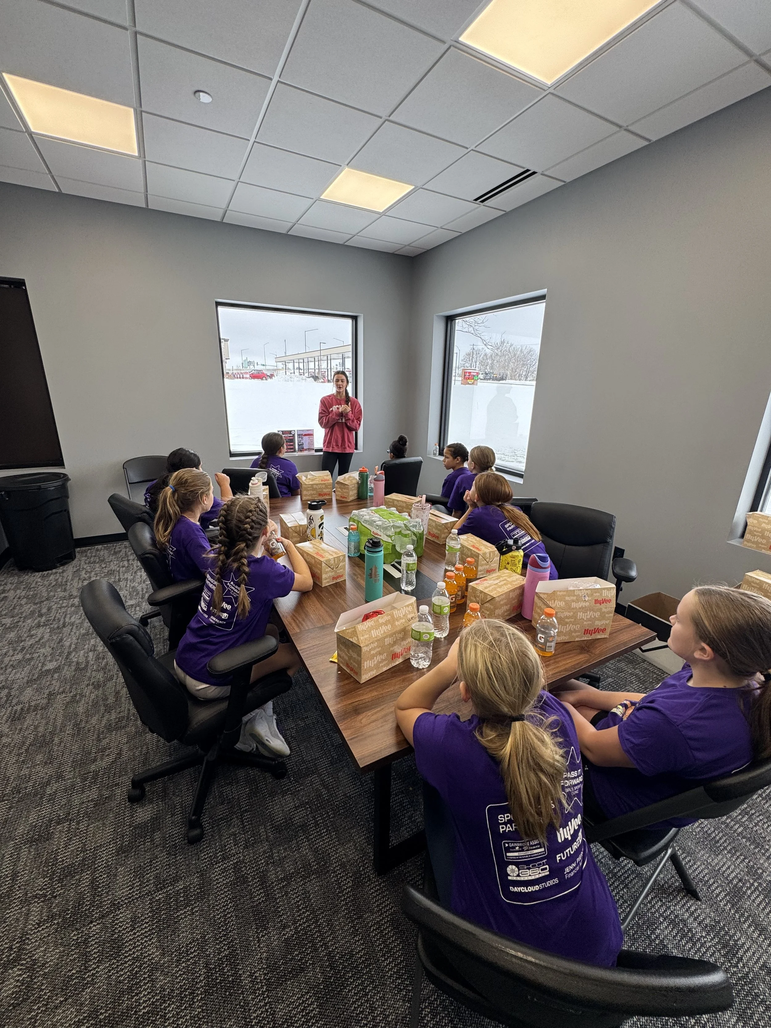 A group of children in purple shirts sitting around a conference table in a room with large windows and a gray ceiling, listening to a woman speaking at the front.
