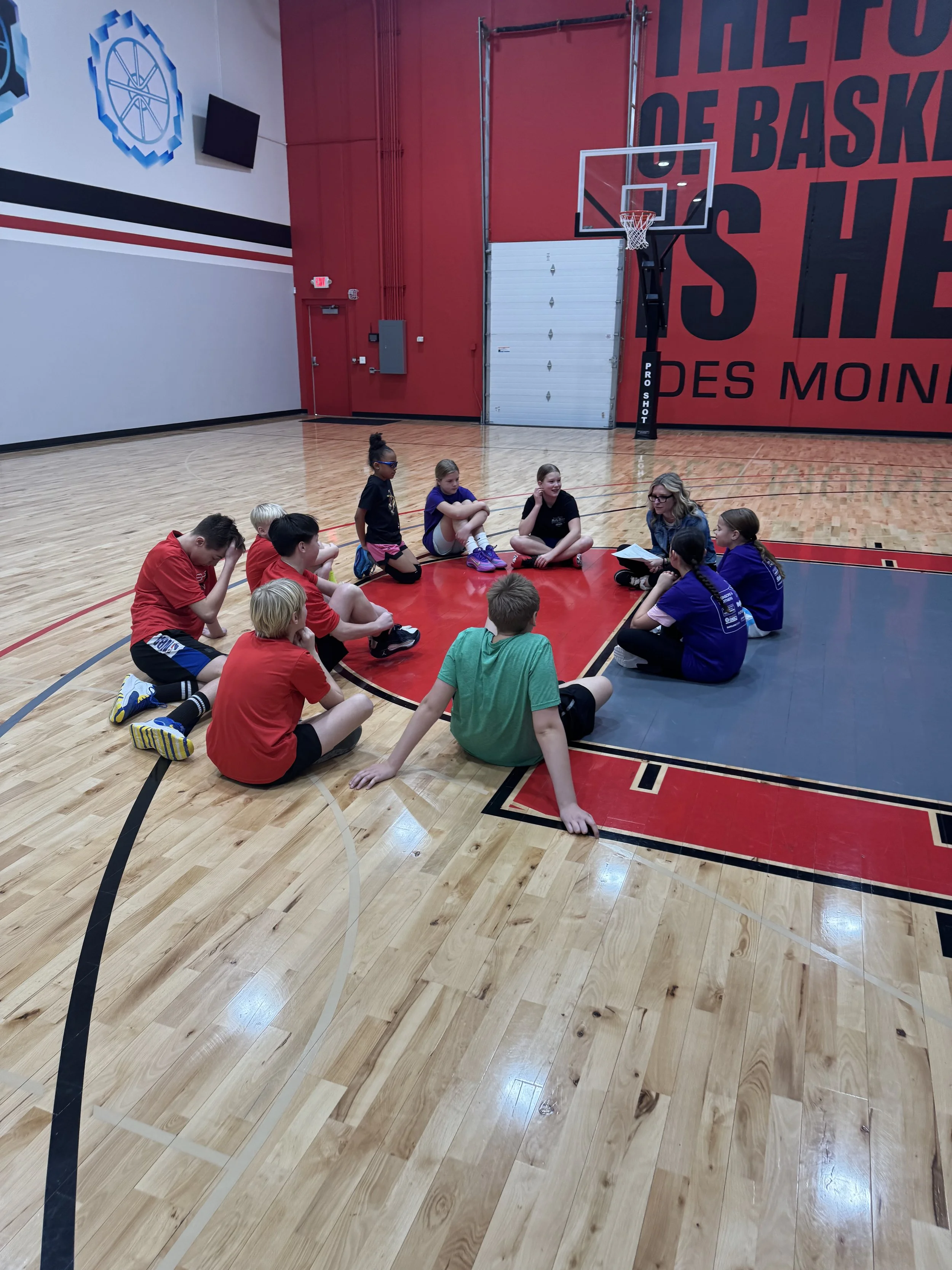 A group of children sitting on the basketball court floor inside a gym, listening to an adult woman with blonde hair and glasses, who is holding a clipboard and speaking to them.