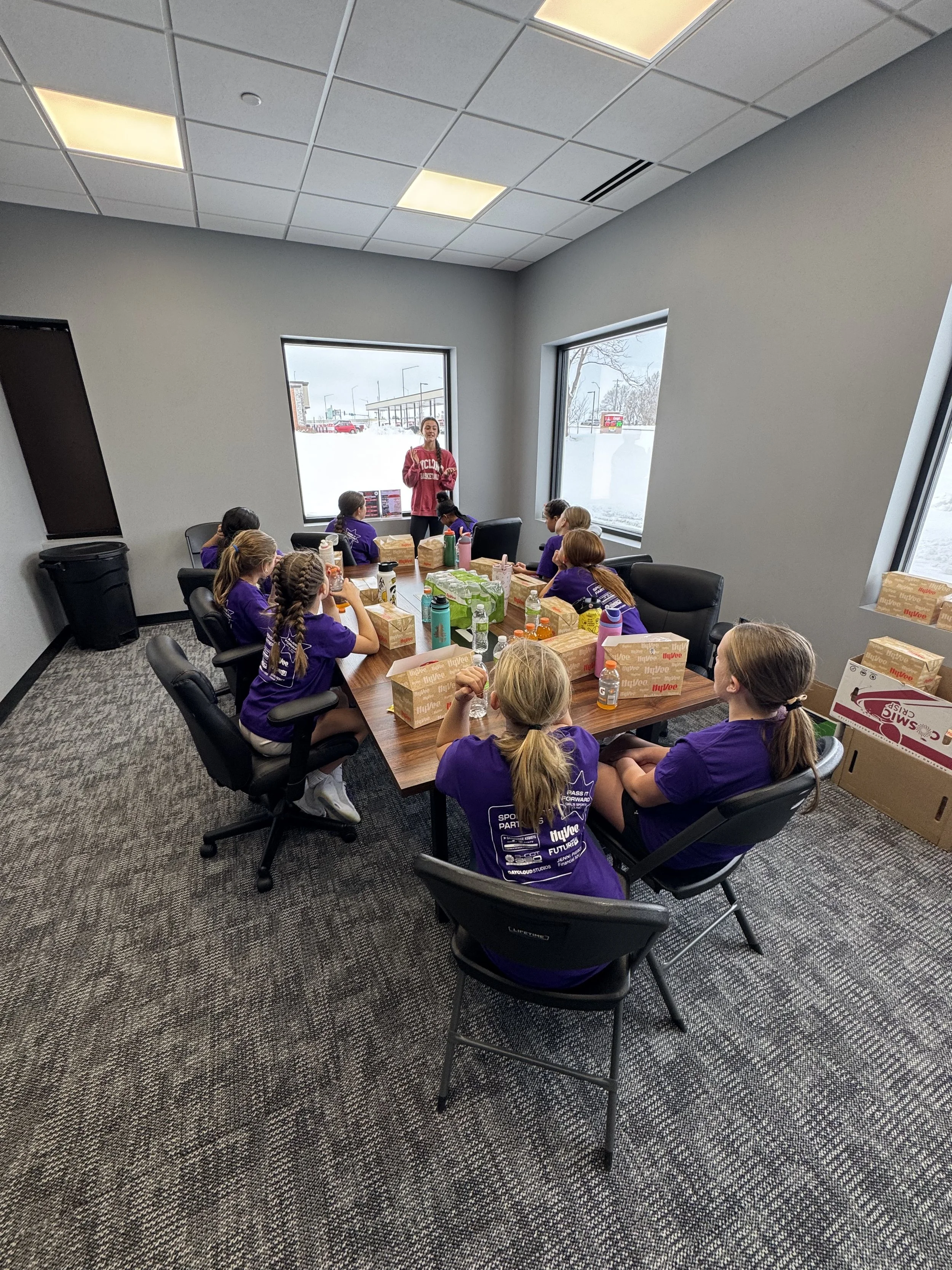 A group of children wearing purple shirts sitting around a conference table with food boxes and drinks, while a woman in a red shirt stands near the windows.