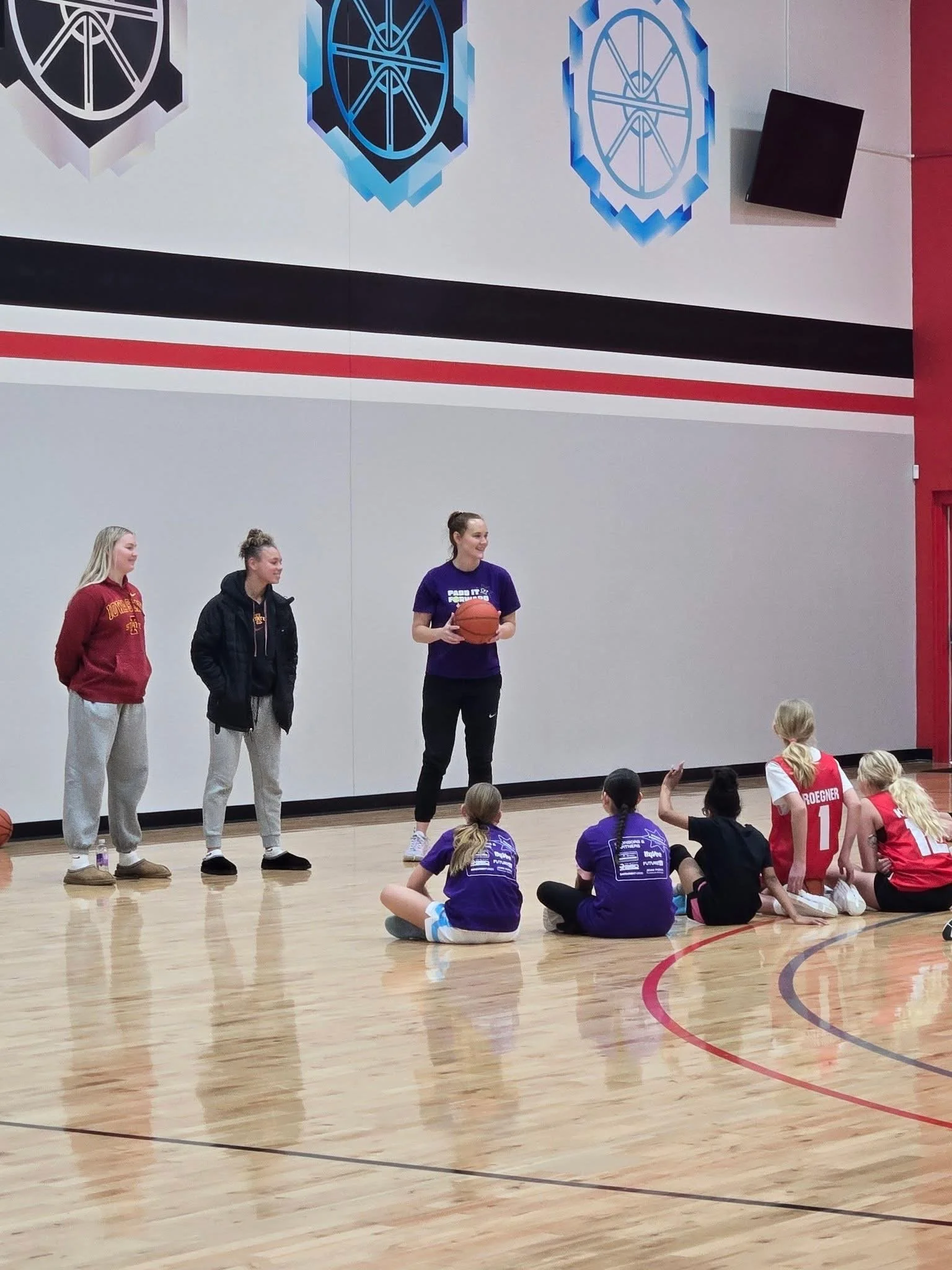 Indoor gymnasium with a basketball court, basketball players sitting on the floor, and three women standing on the court. One woman is holding a basketball.