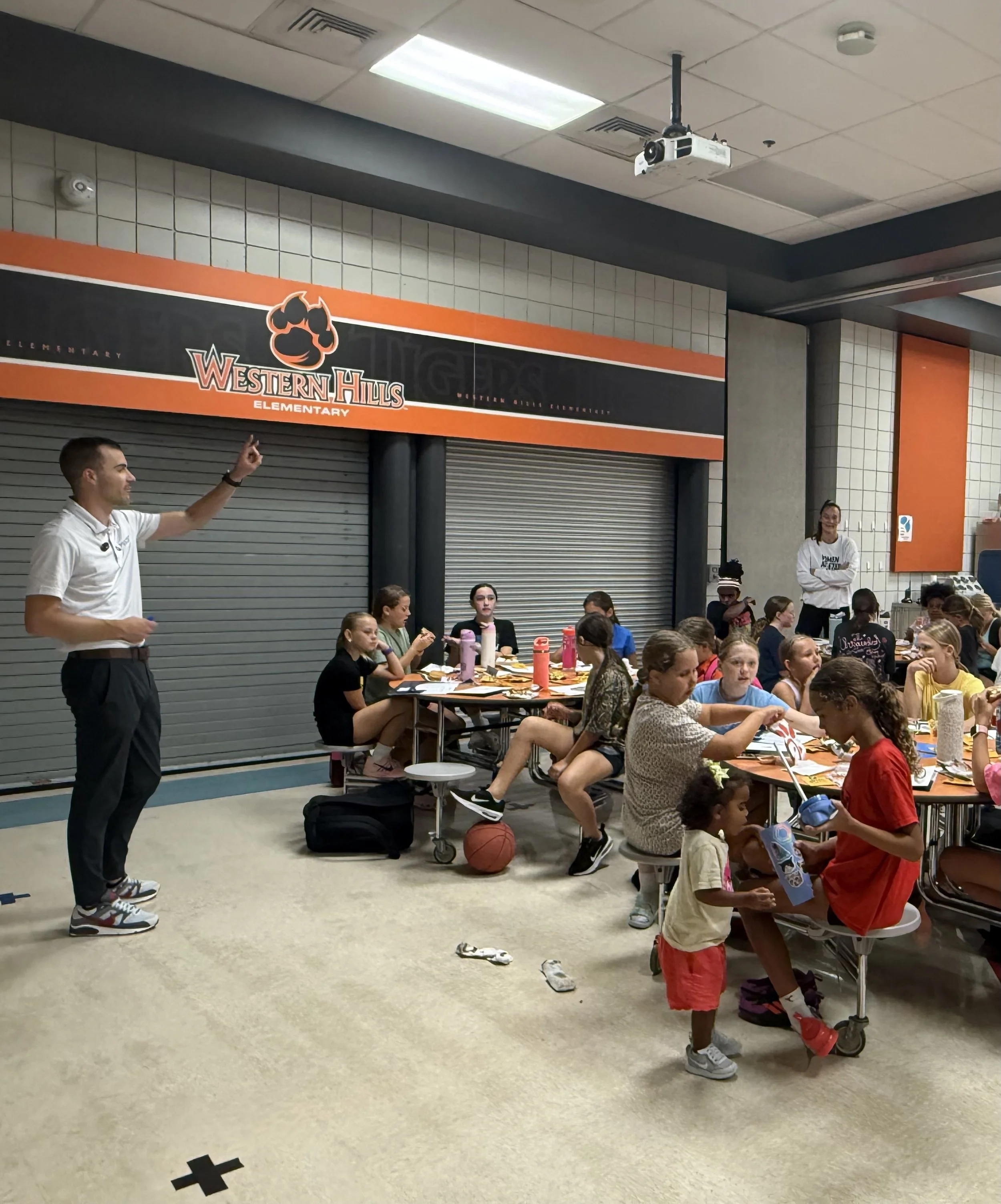 A group of elementary school children and two adults are gathered in a school cafeteria for a meal or event. Some children sit on stools at a long table, while others, including two girls in red shirts, are standing and handing out items. The background features a school sign for Western Hills Elementary and a closed gray shutter.