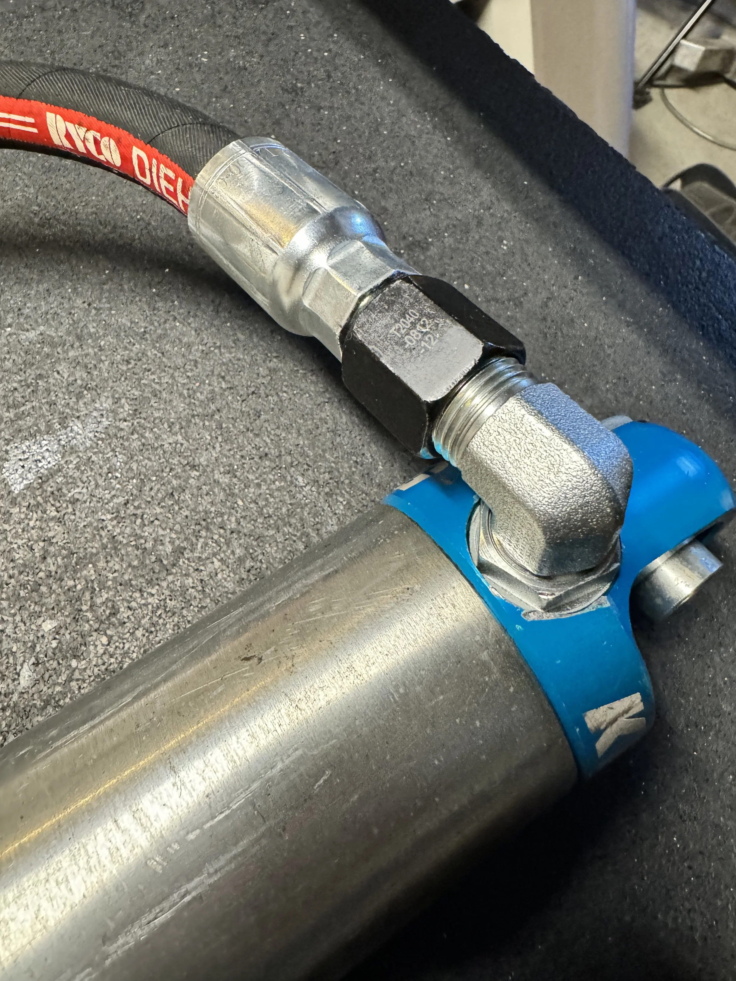 Close-up of a large metal pipe connected to a valve with blue fittings, a red hose with branding, and various metal fittings and pipes on a gray textured surface.