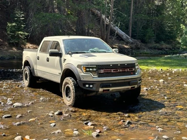 A beige pickup truck is parked in a shallow stream surrounded by trees and forest.