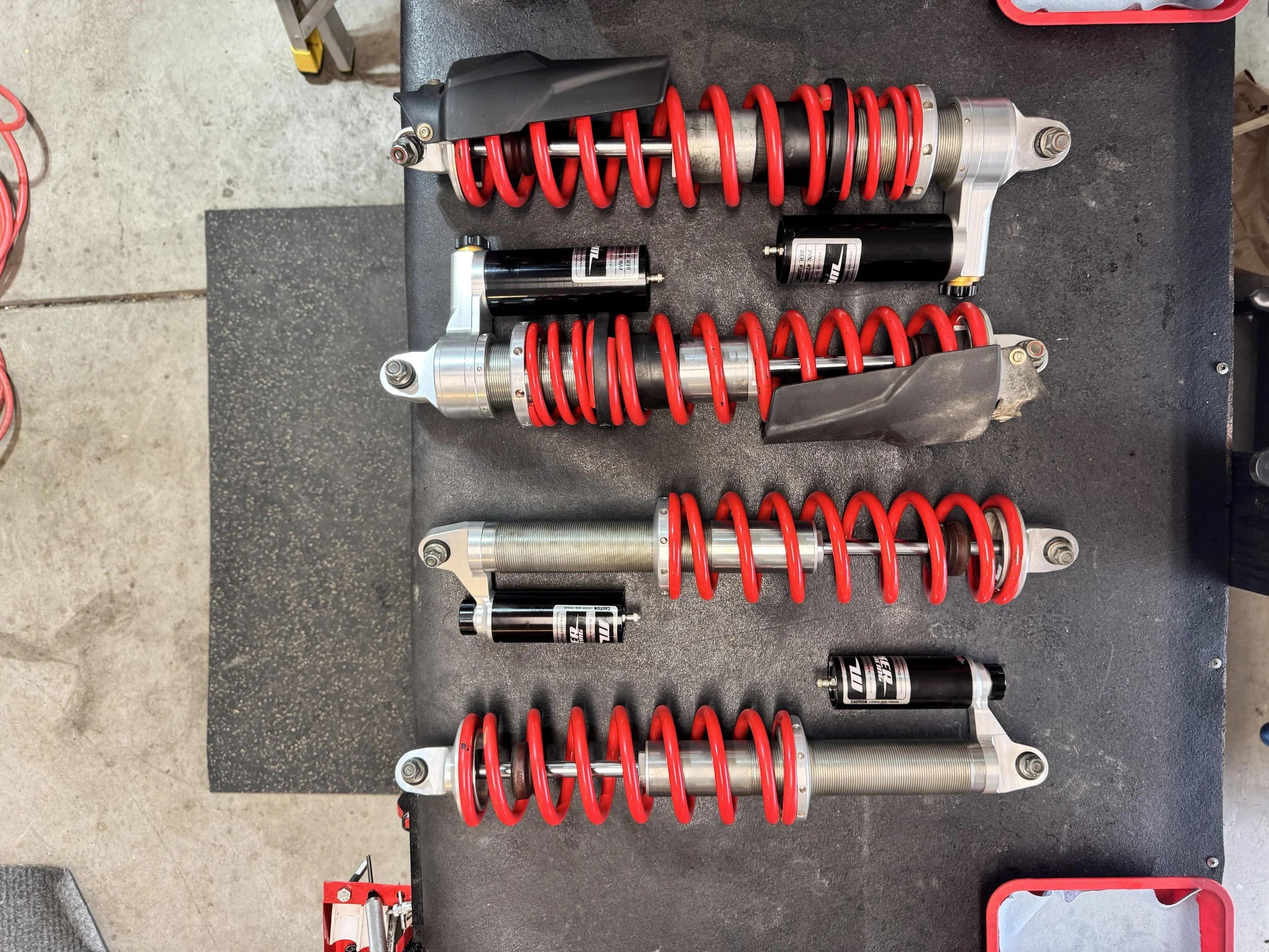 A set of new motorcycle suspension forks with red springs, shock absorbers, and associated hardware laid out on a black workbench.