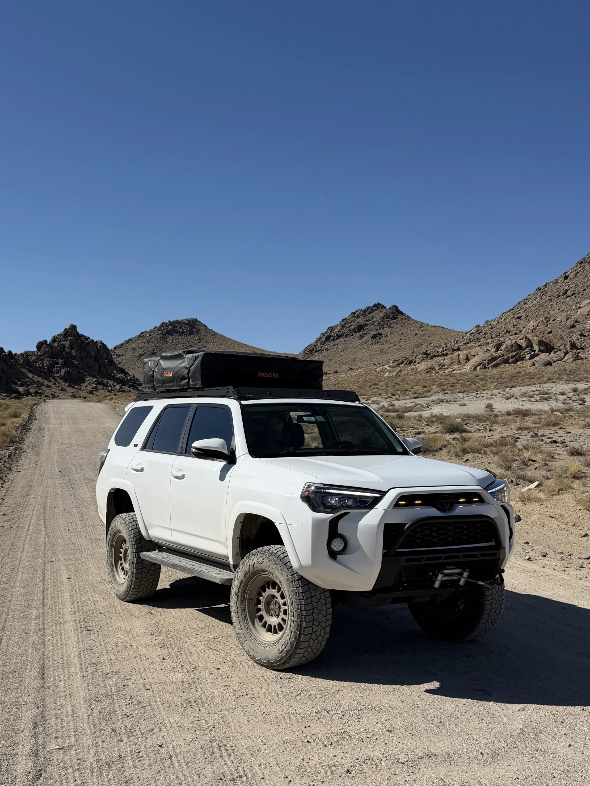 A white off-road vehicle driving on a dirt road in a desert landscape with rocky hills and clear blue sky.