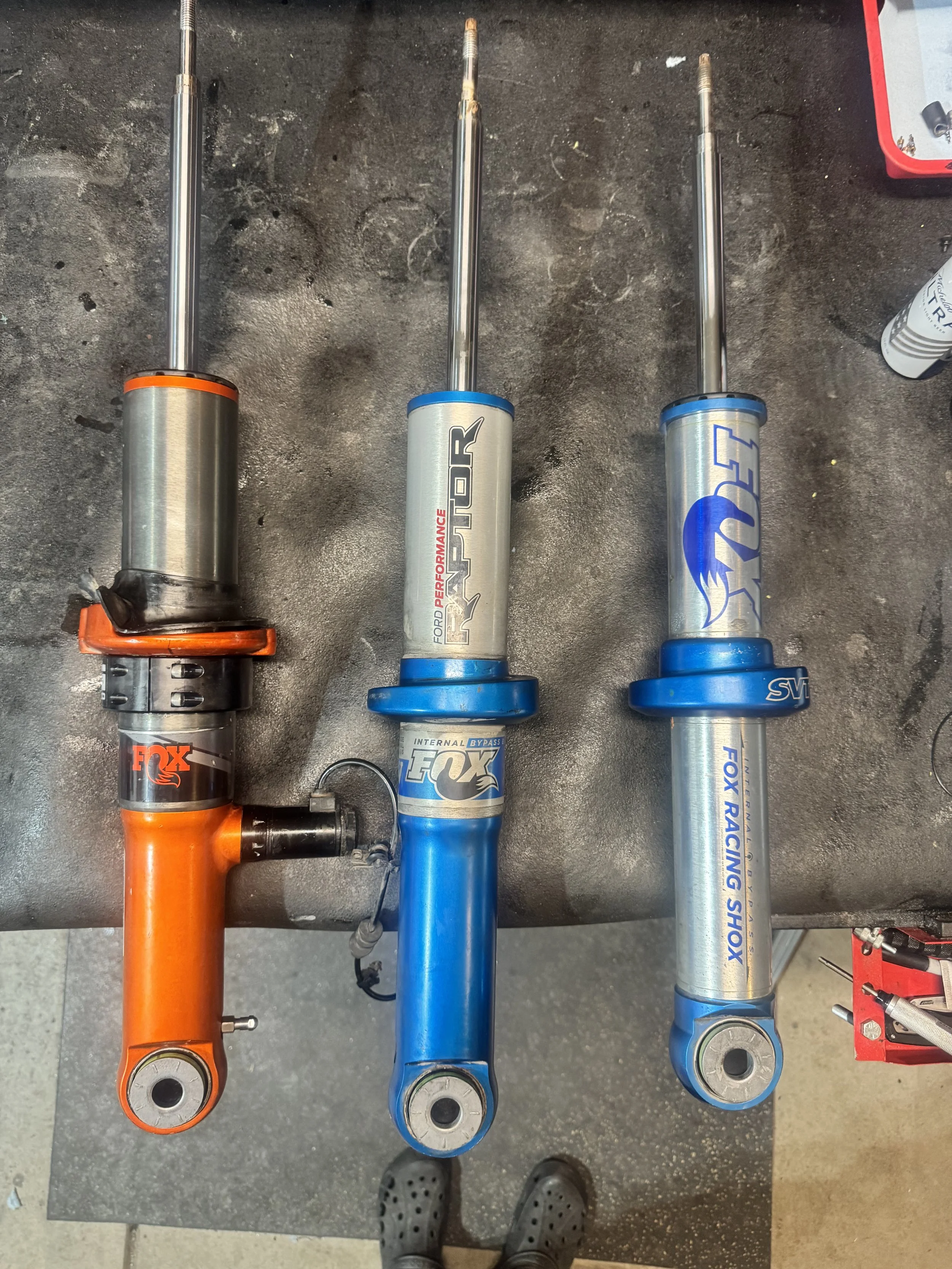 Three motorcycle front forks placed on a workbench, with the person wearing Crocs shoes visible in the background.