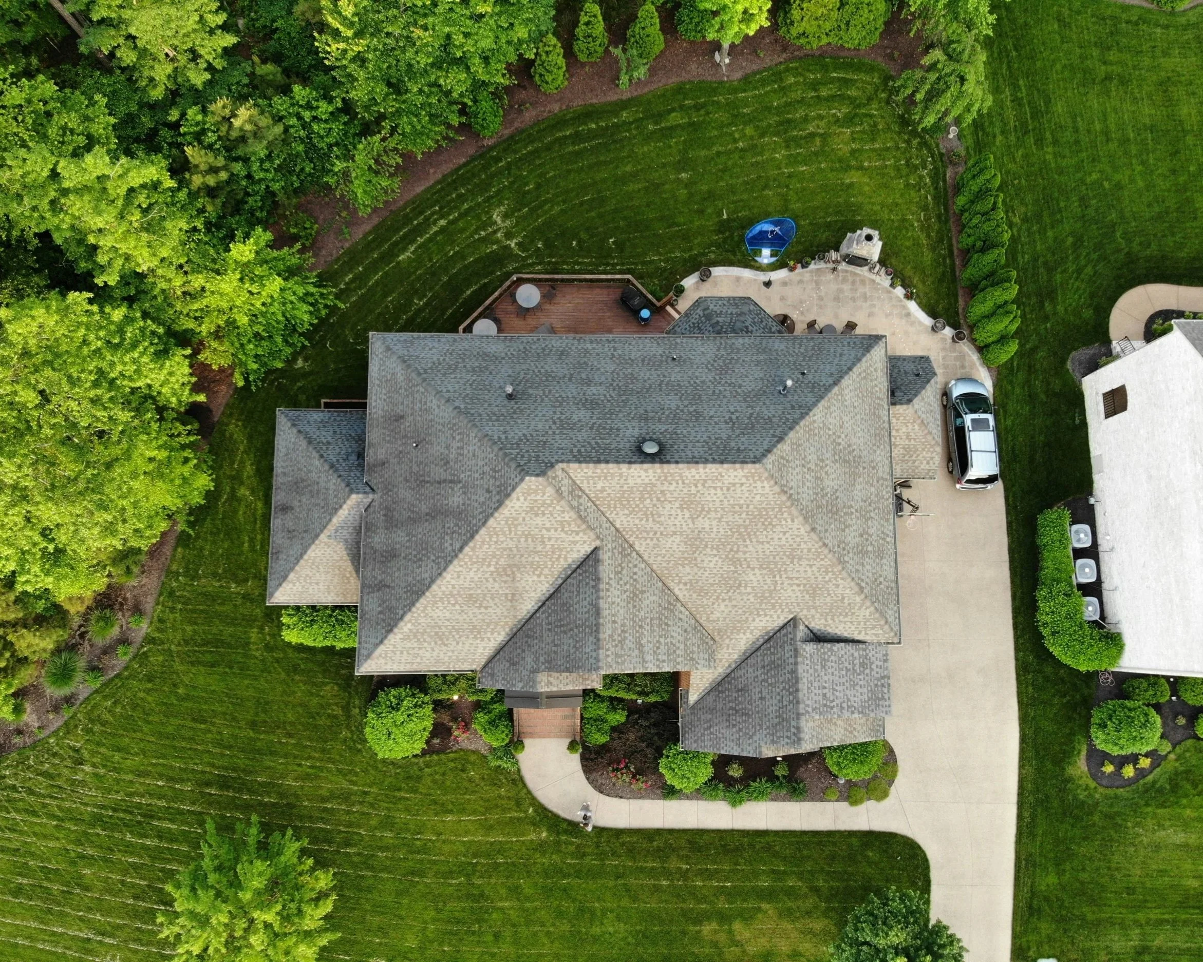 An aerial view of a suburban backyard showing a house with a gray roof, a patio with outdoor furniture, a grassy lawn, and lush green trees along the edges.